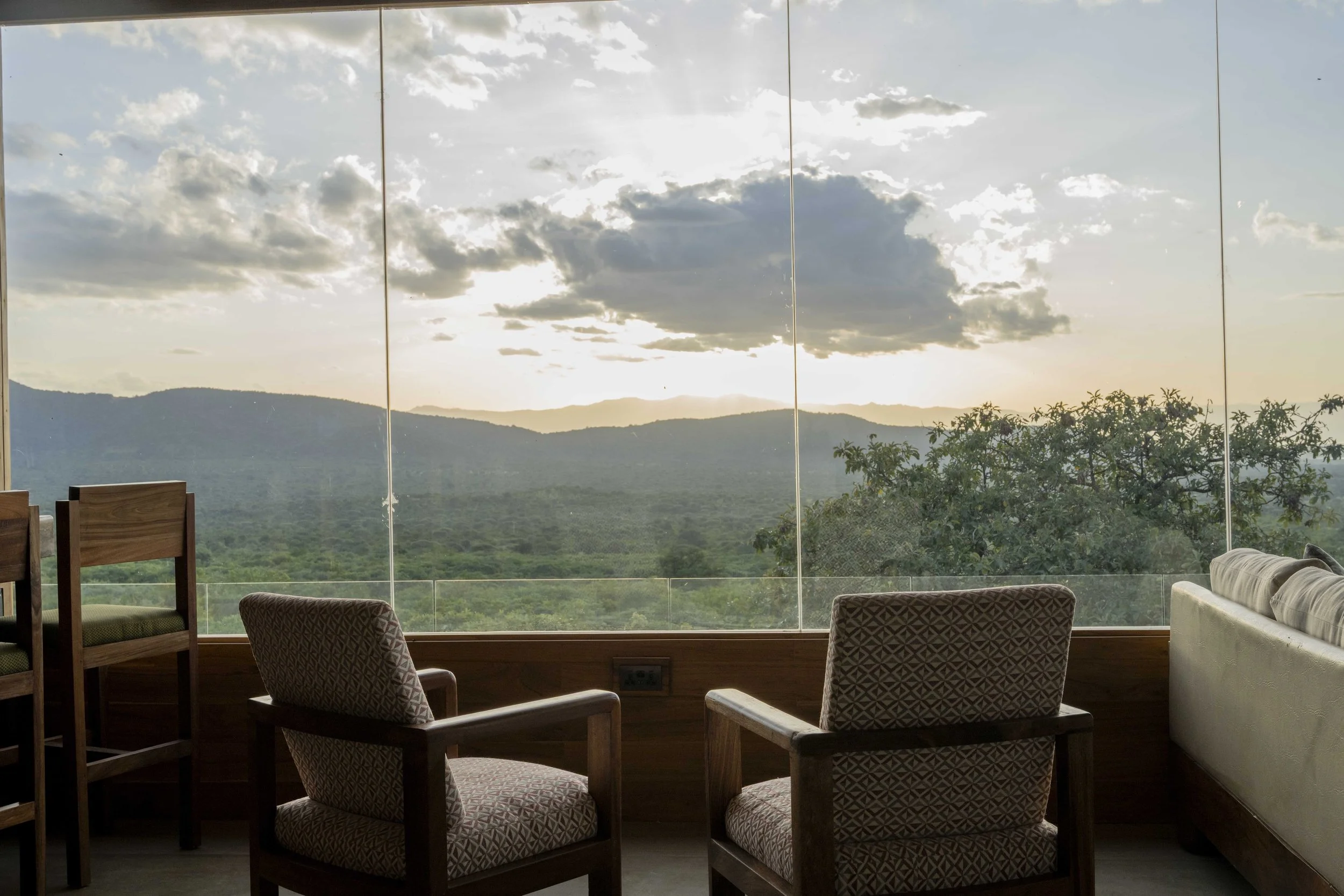 Interior of a room with a large glass window showing a scenic mountain view and clouds in the sky, with chairs and a sofa in the foreground.