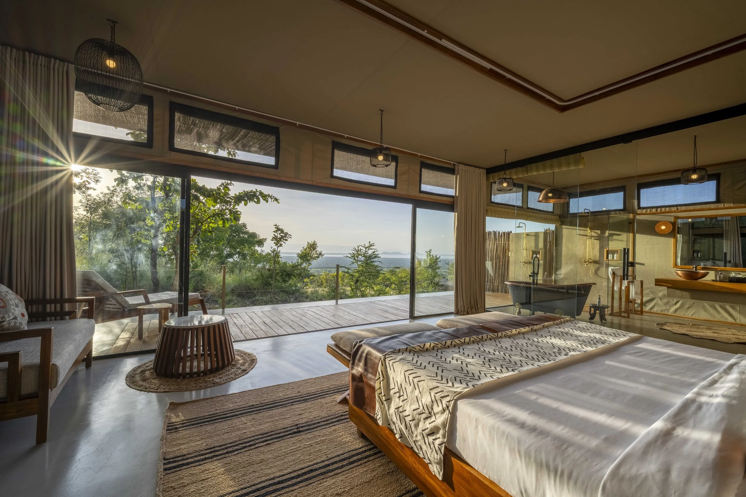A modern bedroom with large glass sliding doors opening to a deck and greenery outside, featuring a bed with patterned bedding, a sitting area with chairs and a small table, and pendant light fixtures hanging from the ceiling.