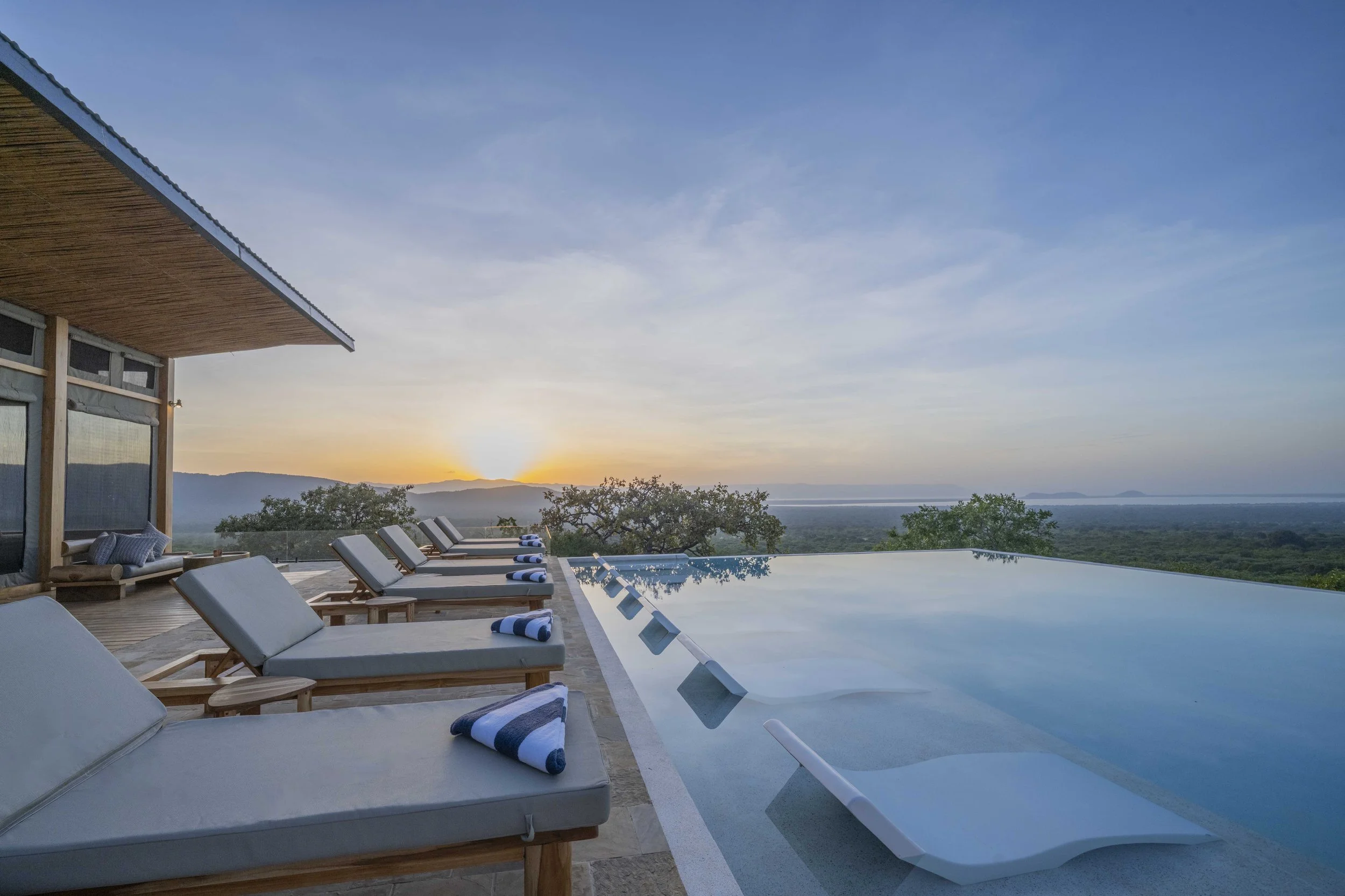 Luxury outdoor pool area with lounge chairs and a view of mountains and a sunset.