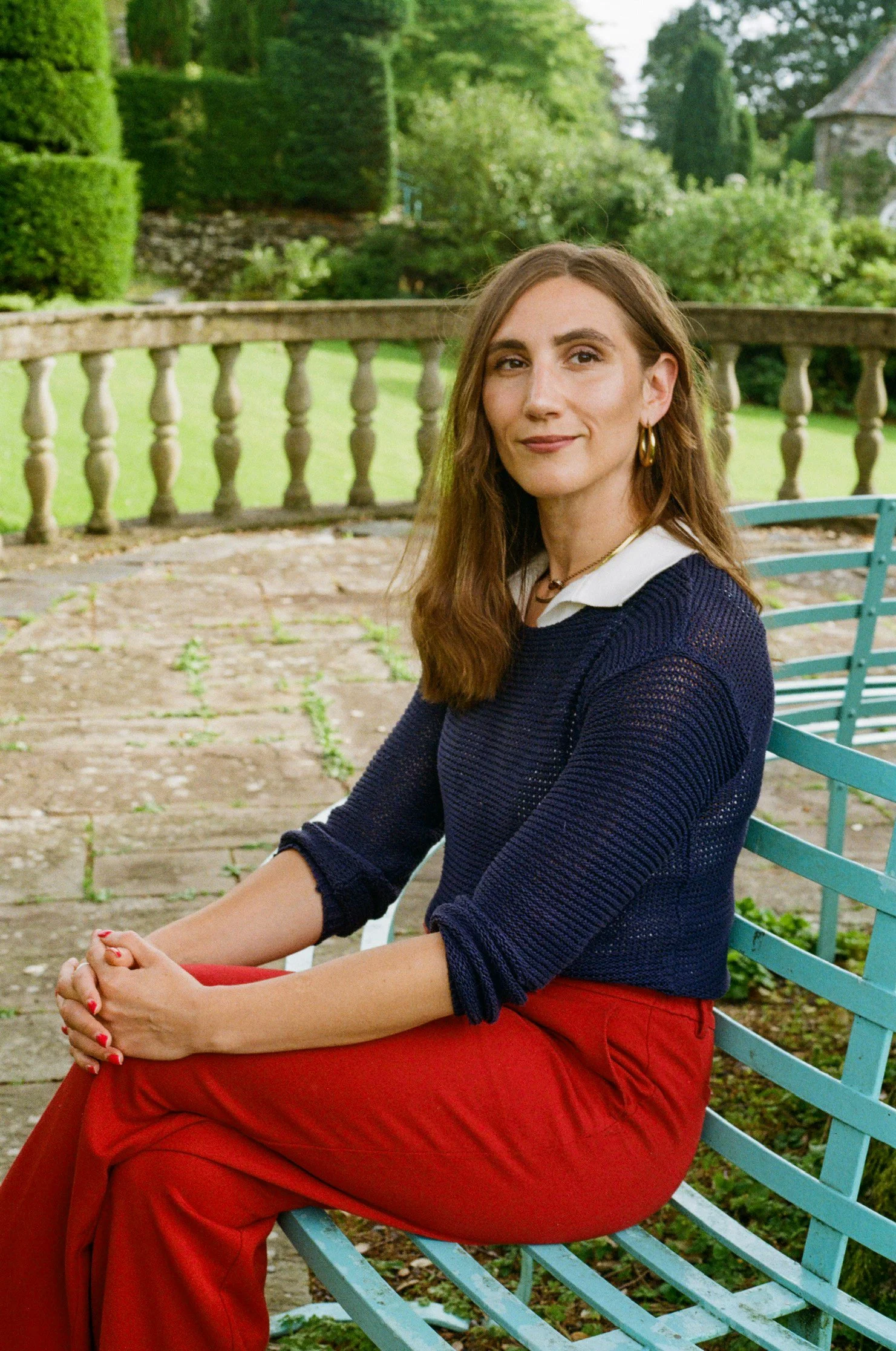 A woman with long brown hair, wearing a navy blue sweater over a white collared shirt, red pants, gold hoop earrings, and layered necklaces, sitting on a light blue metal bench outdoors with greenery and a stone railing in the background.