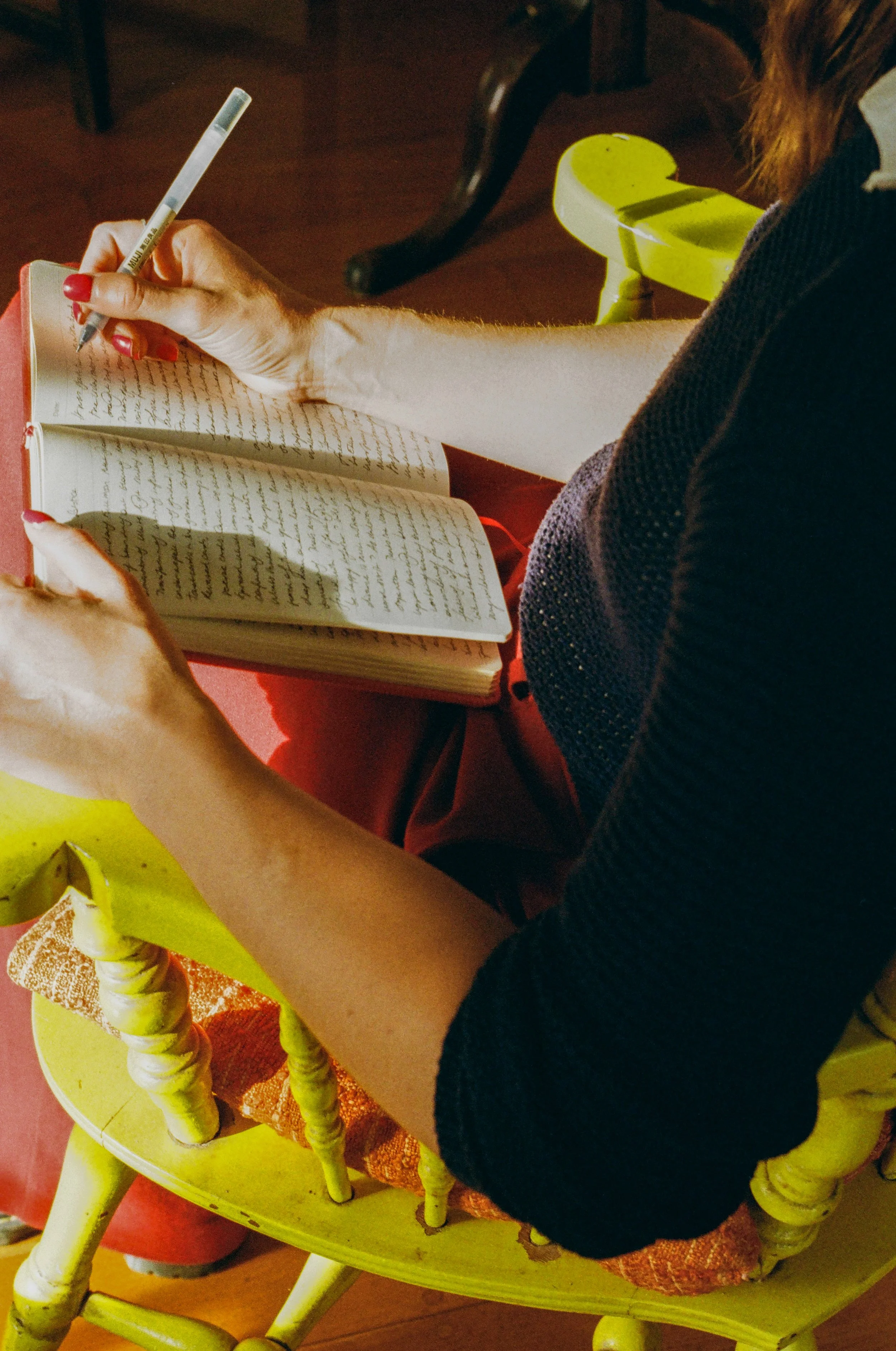 Person sitting on a yellow chair, writing in a notebook with a pen, with a wooden floor and part of a table visible.
