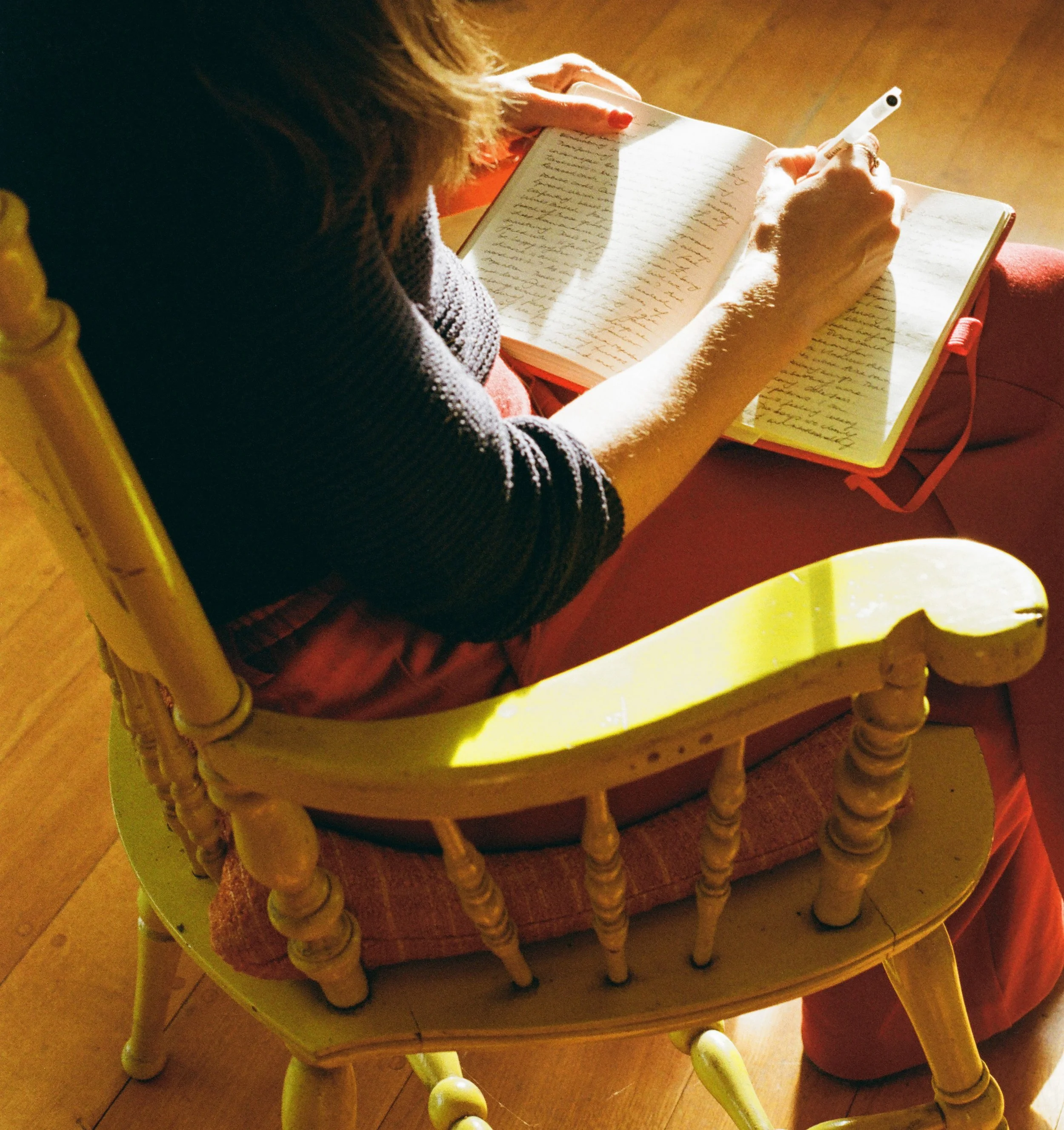 A person sitting on a yellow wooden chair, writing in a notebook with a pen. The person has light skin and is wearing a gray top and reddish pants. The scene is lit by natural sunlight.