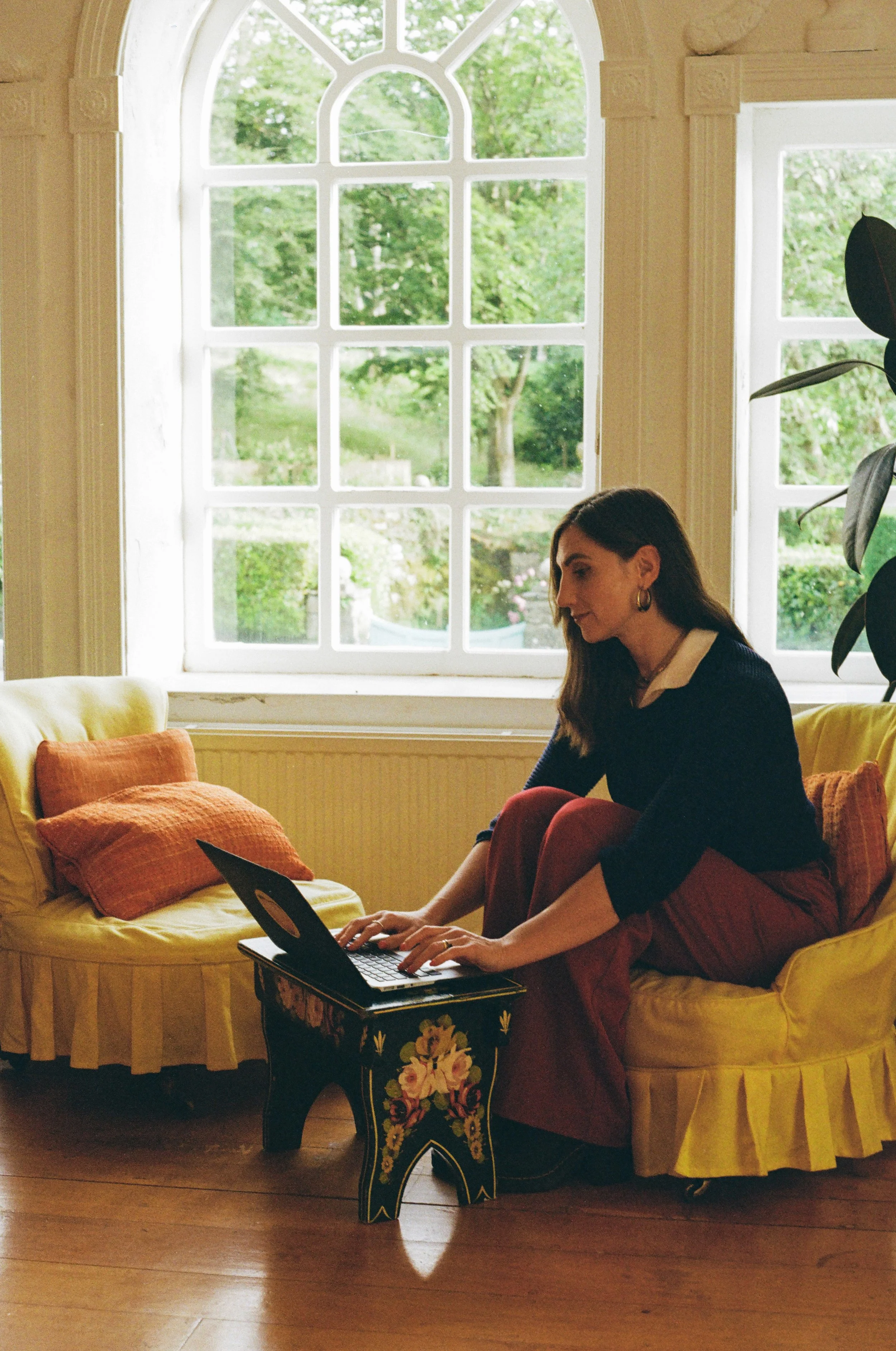 A woman sitting on a yellow sofa using a laptop in a room with large windows and green trees outside.