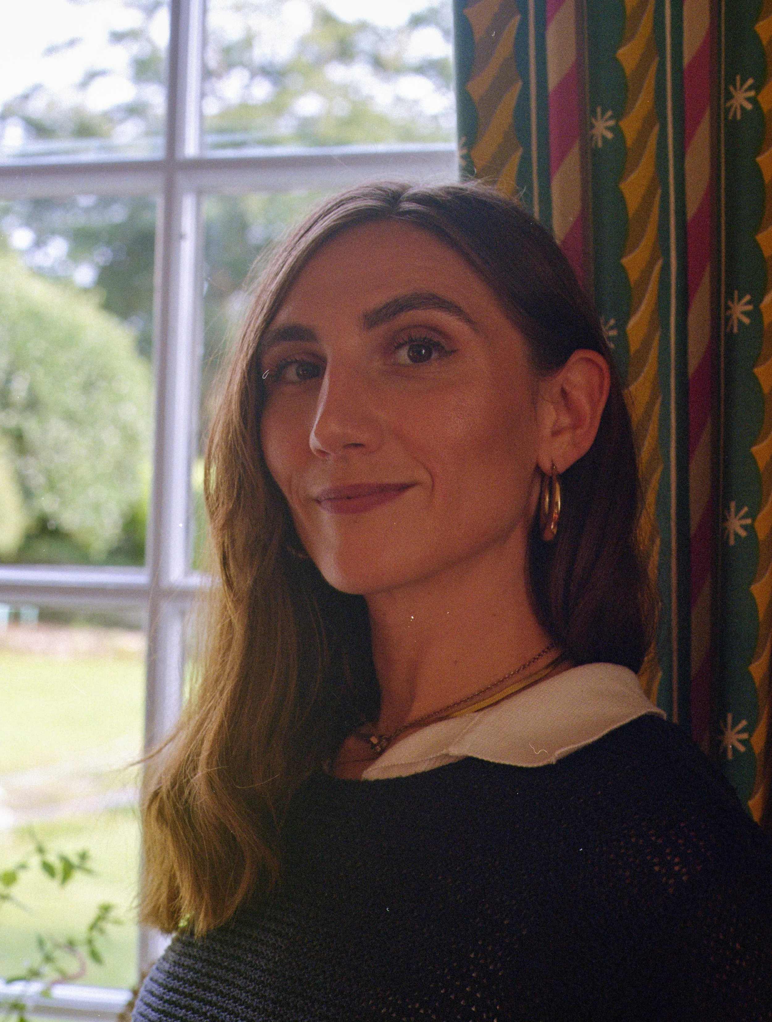 A woman with long brown hair and hoop earrings standing near a window with a patterned curtain, smiling slightly.