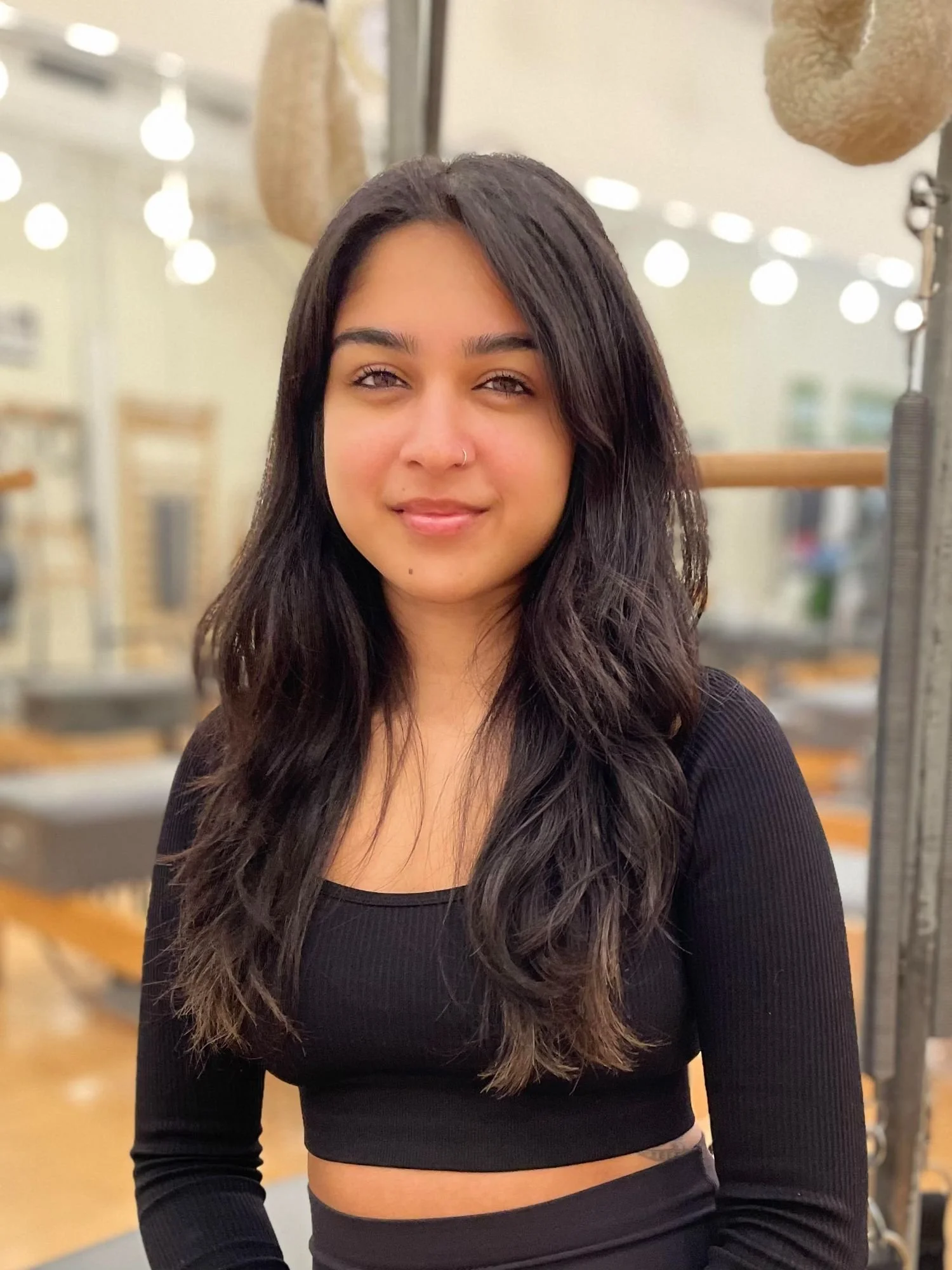 A young woman with long dark wavy hair, wearing a black top, standing in a Pilates studio with blurred background and warm lighting.