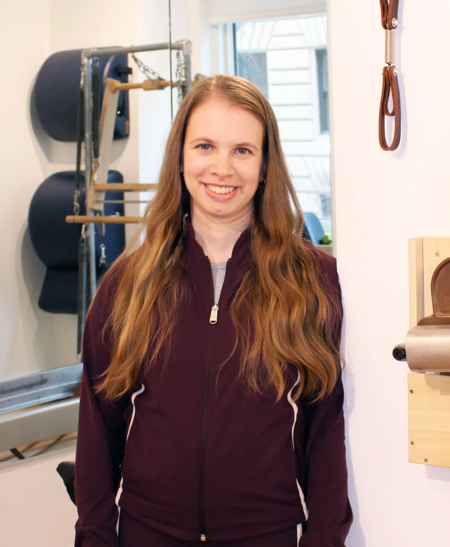 A woman with long wavy hair smiling in a Pilates studio, standing near Pilates equipment like a wall-mounted rack with black pads and wooden bars.