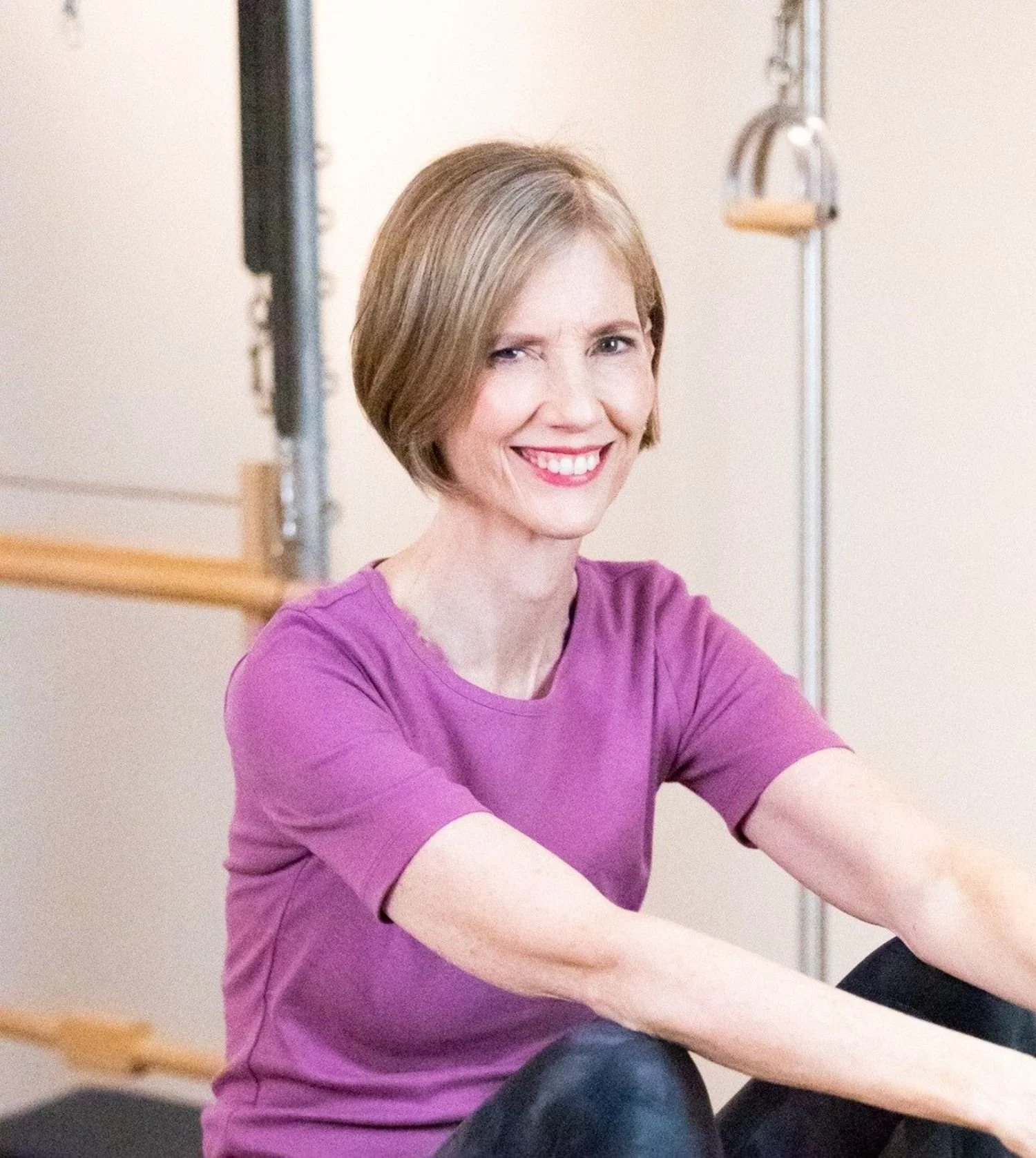 A woman with short brown hair wearing a purple shirt, smiling and sitting in a room with Pilates equipment.