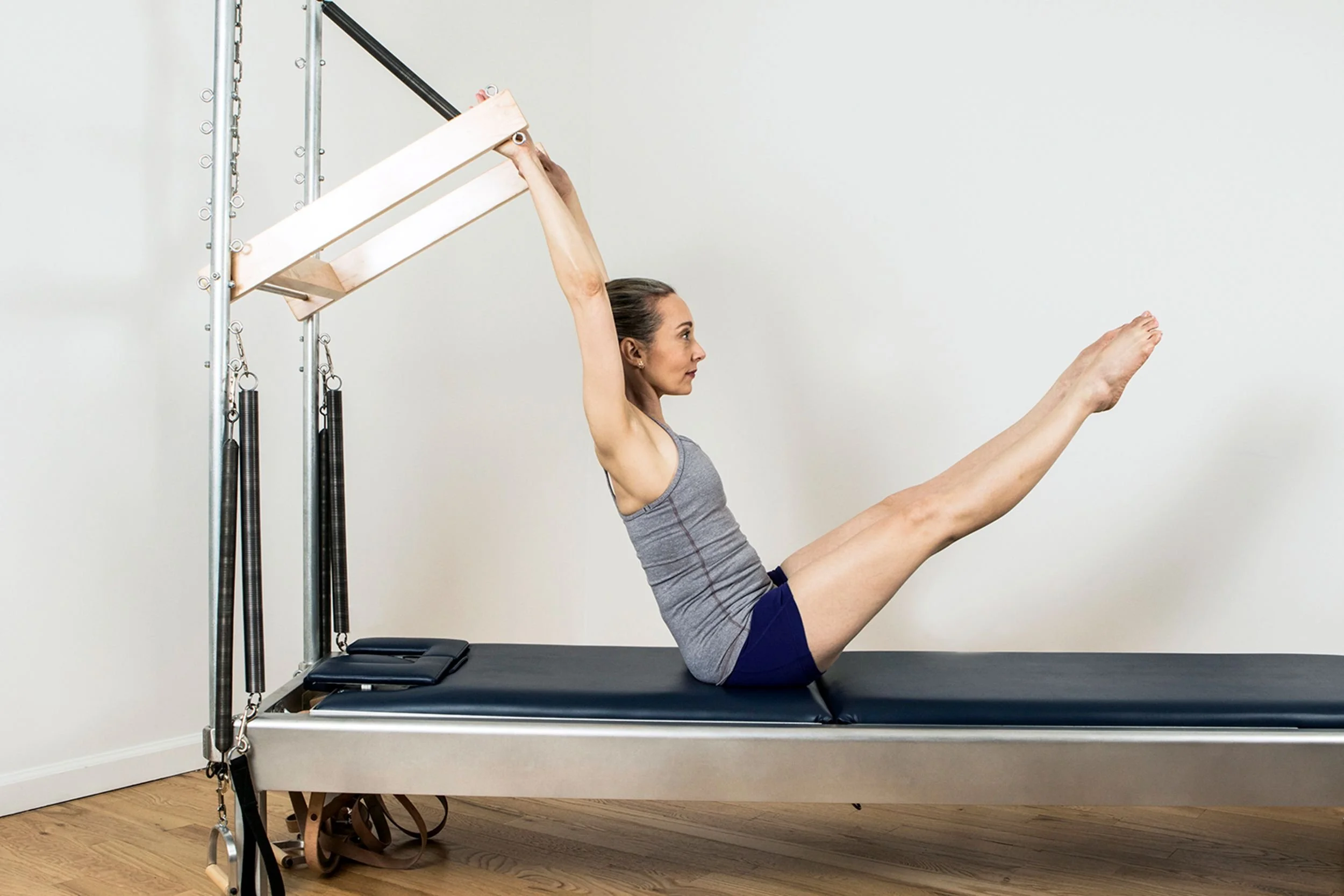 Woman exercising on a Pilates reformer machine, lying on her back with legs extended, holding a wooden bar overhead.