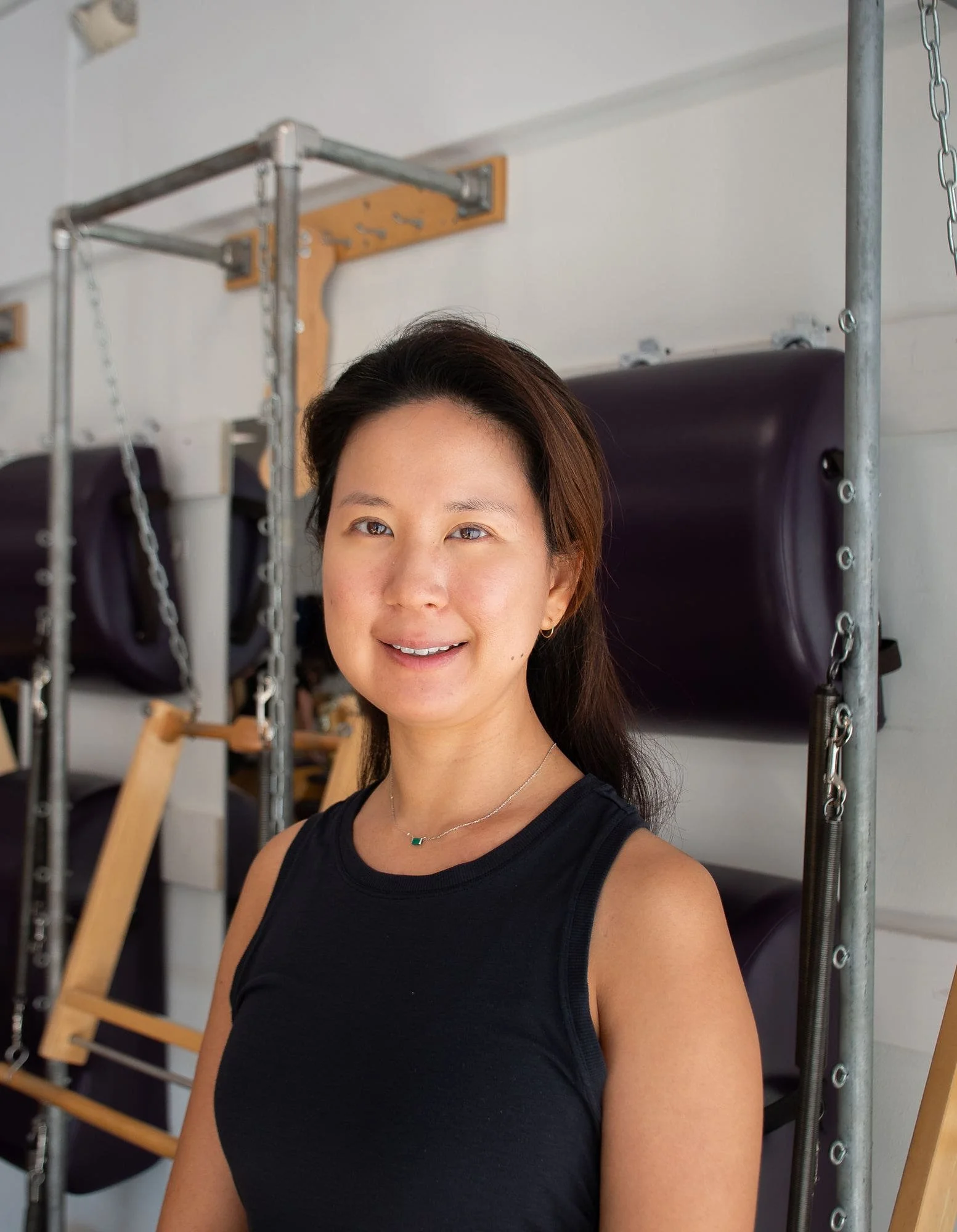 A woman with long dark hair, wearing a black top, standing in a Pilates studio.