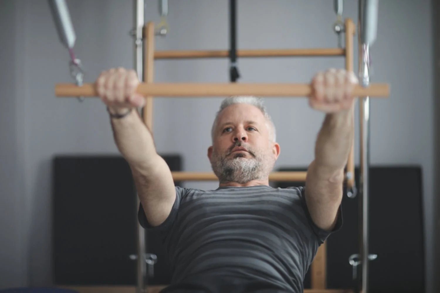 A middle-aged man with gray hair and beard doing a workout on a wooden pull-up bar in a gym.