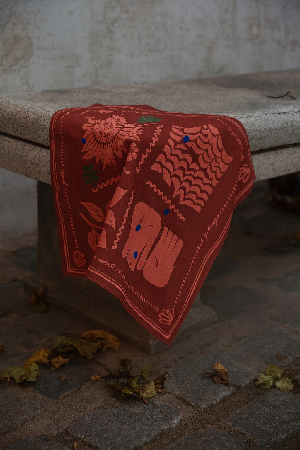 A red bandana with floral and geometric patterns draped over the corner of a gray stone bench, with scattered leaves on a cobblestone ground.