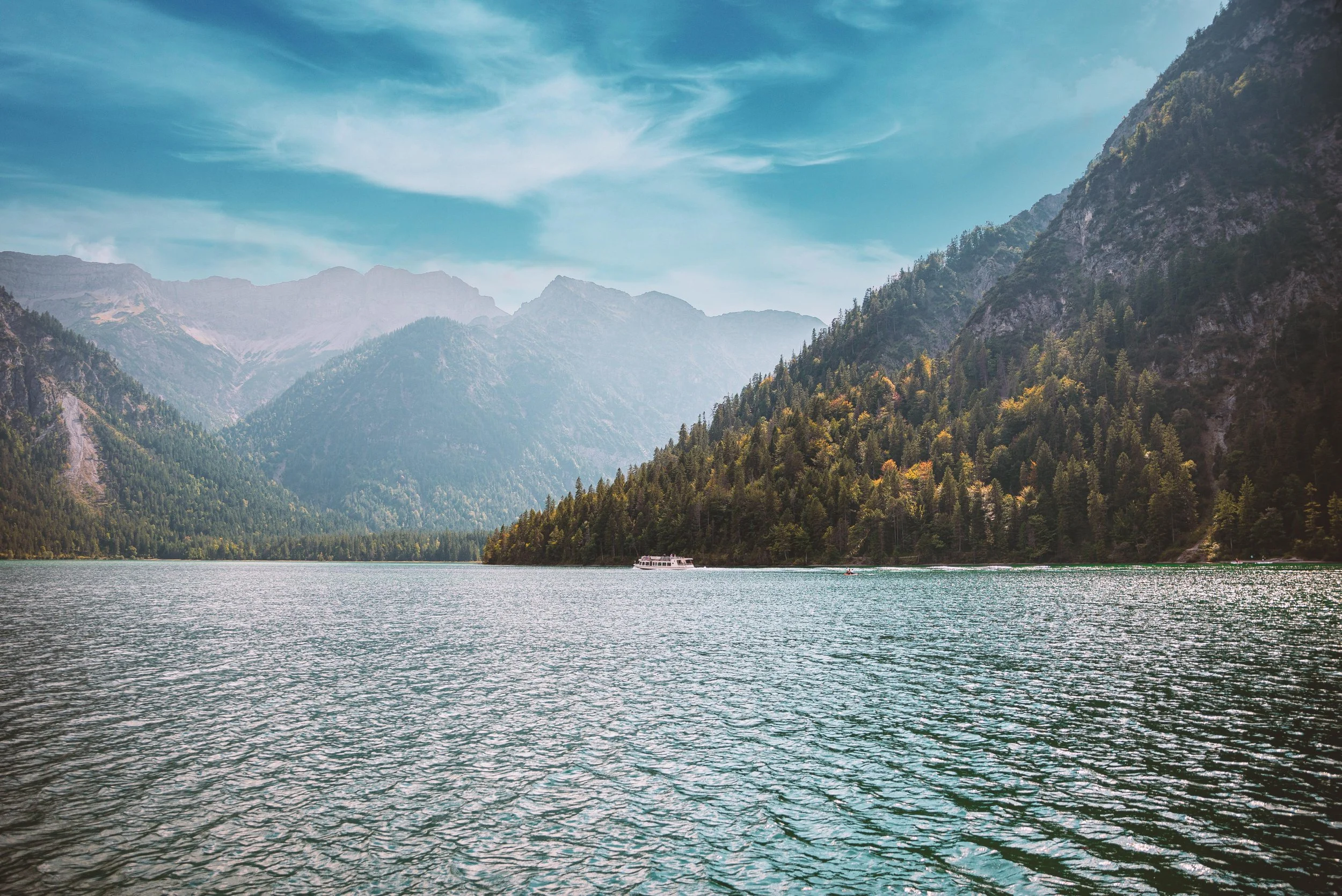 A scenic view of a lake surrounded by towering green mountains under a partly cloudy sky, with a boat on the water.