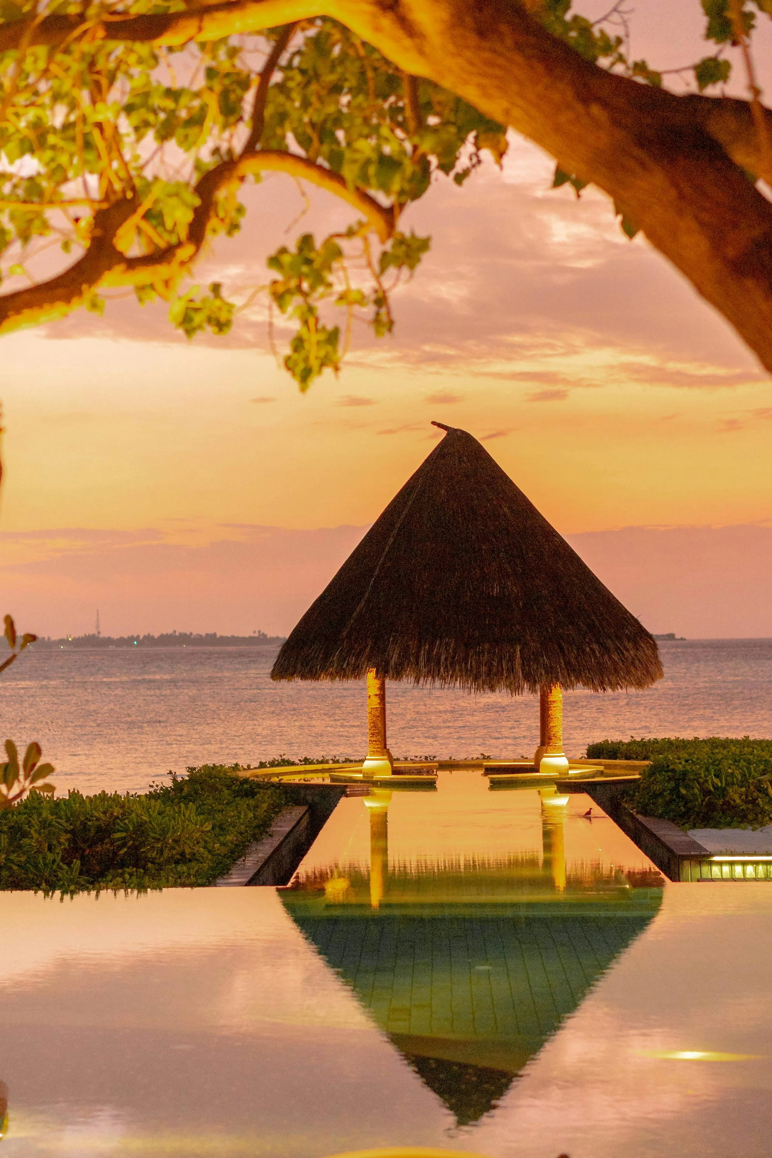 Sunset over a cloudy sky with a thatched-roof pavilion and a reflecting pool on a coastal resort.