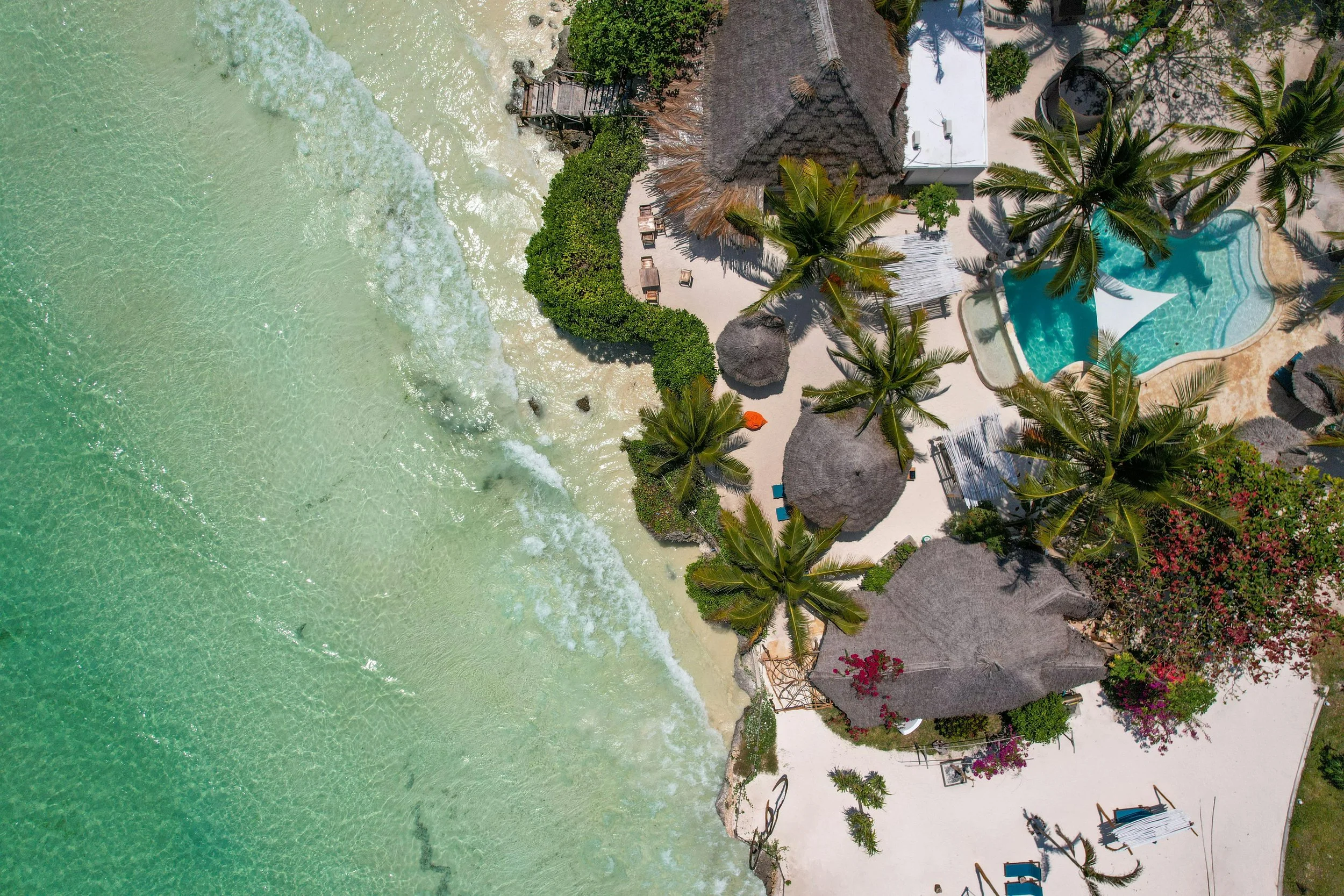 Aerial view of a tropical beach resort with palm trees, thatched-roof huts, a swimming pool, and lounge chairs along the sandy shore.