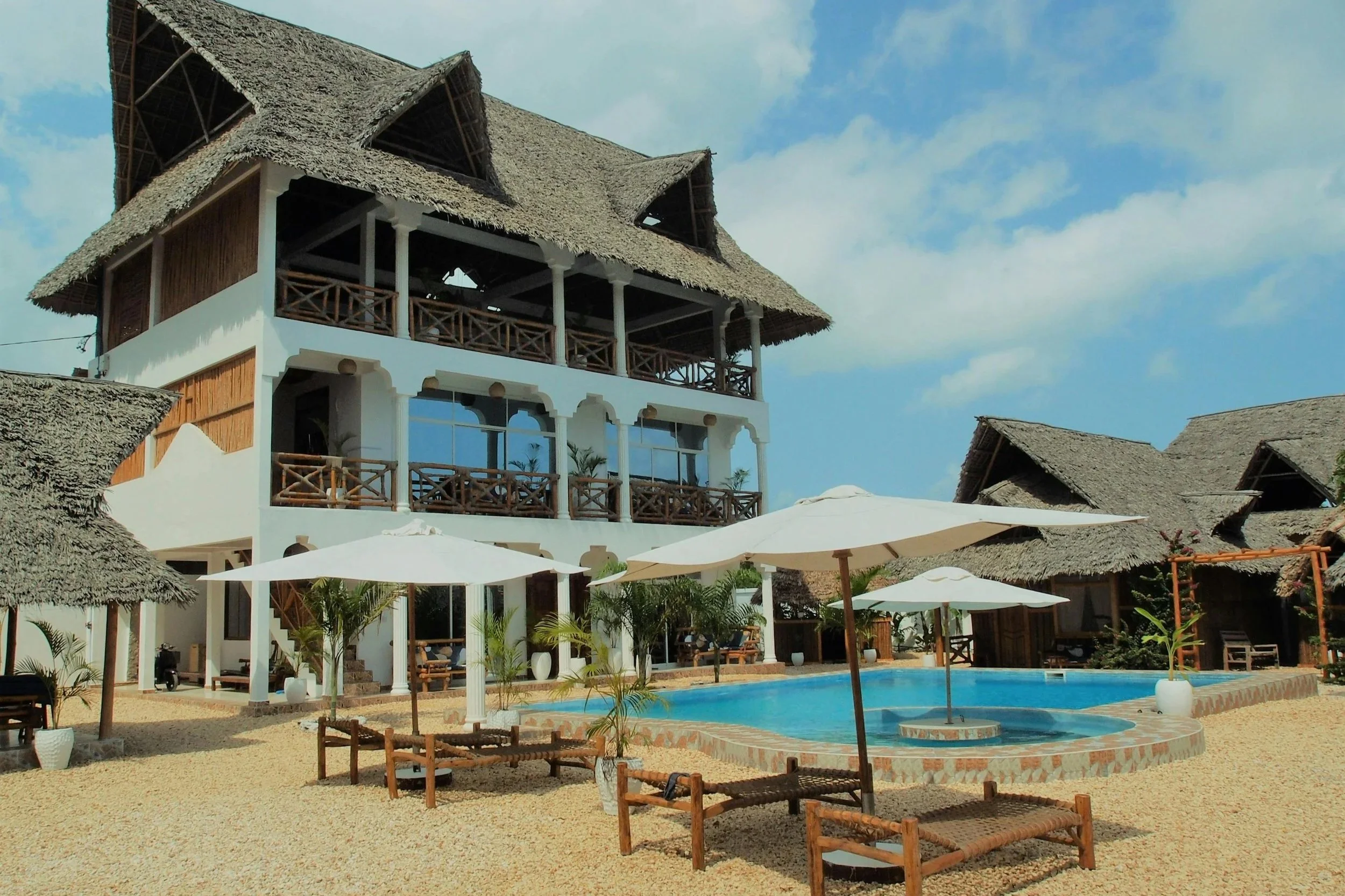 Resort with a swimming pool, sun umbrellas, lounge chairs, and a multi-story building with wooden balconies and thatched roofs.