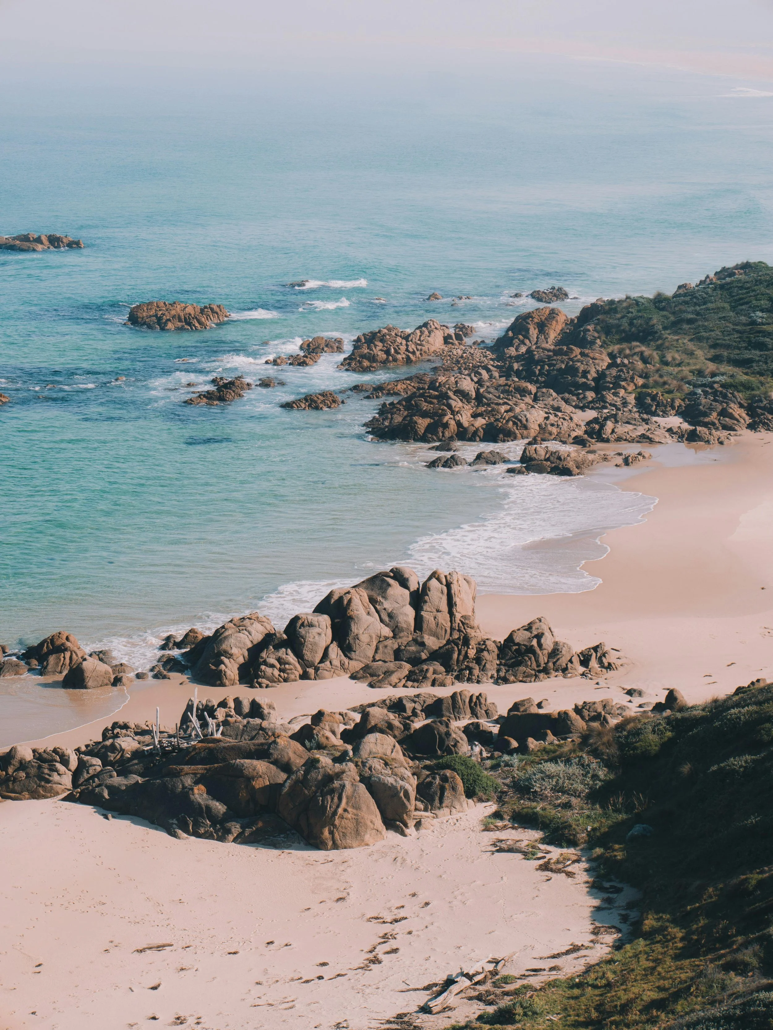 A scenic view of a rocky coastline with turquoise ocean waters, sandy beach, and green hillside.