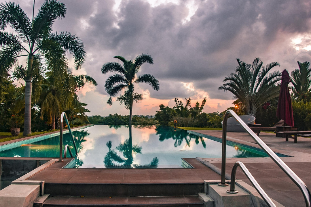 Outdoor swimming pool with palm trees around it at sunset or dusk.