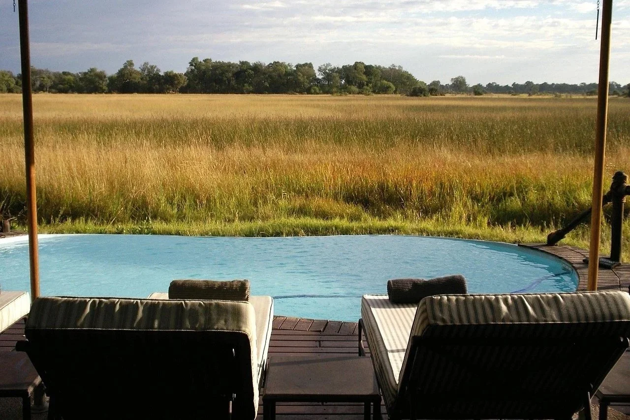 Poolside area with lounge chairs and an umbrella overlooking a grassy field with trees in the distance under a cloudy sky.