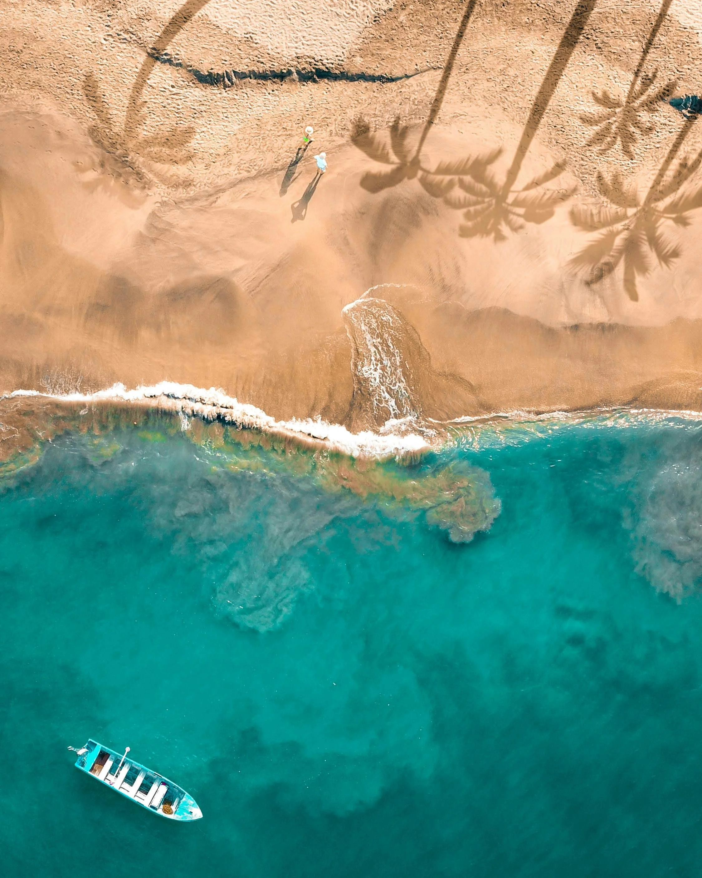 An aerial view of a beach showing two people walking on the sand near the water, with palm trees casting shadows. A boat is floating in the turquoise ocean close to the shore.