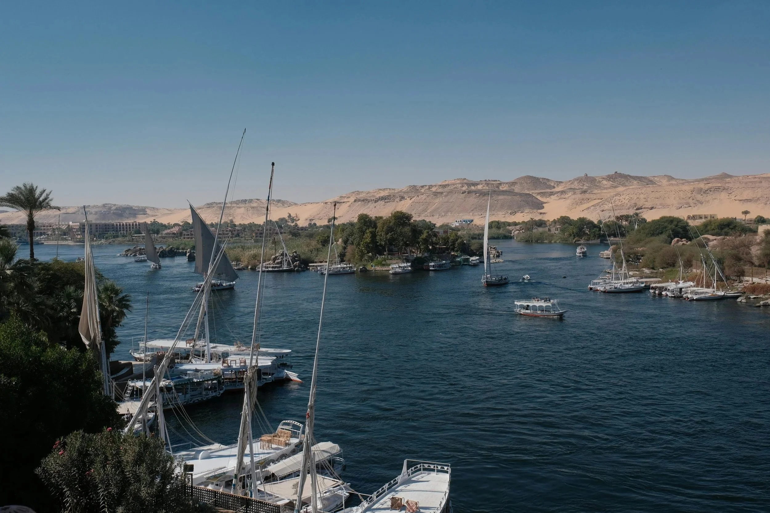 A river scene with boats and sailboats docked along the shore, surrounded by trees and desert hills in the background.