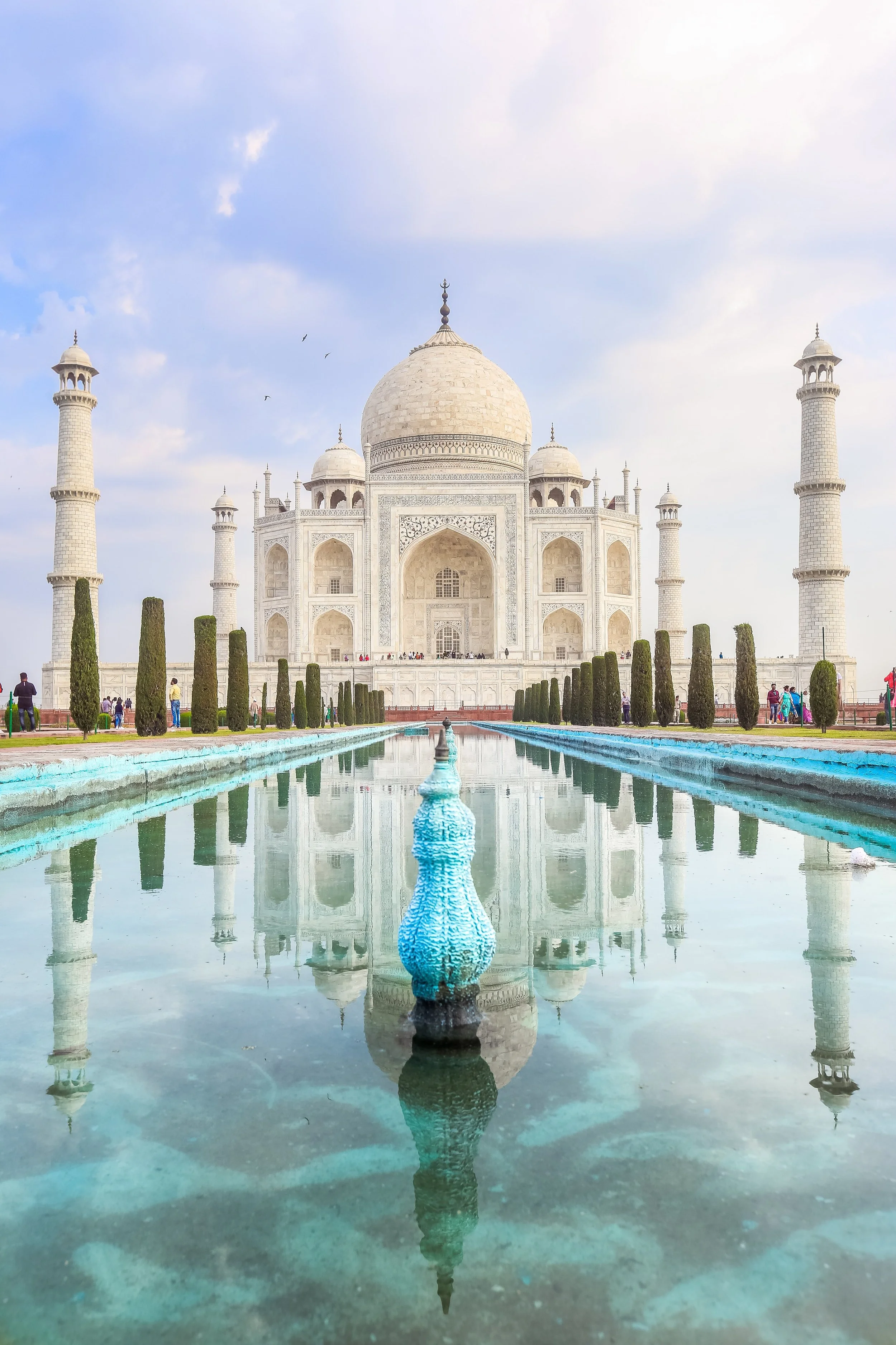 The Taj Mahal, a white marble mausoleum in India, with a reflecting pool in the foreground and people walking around.