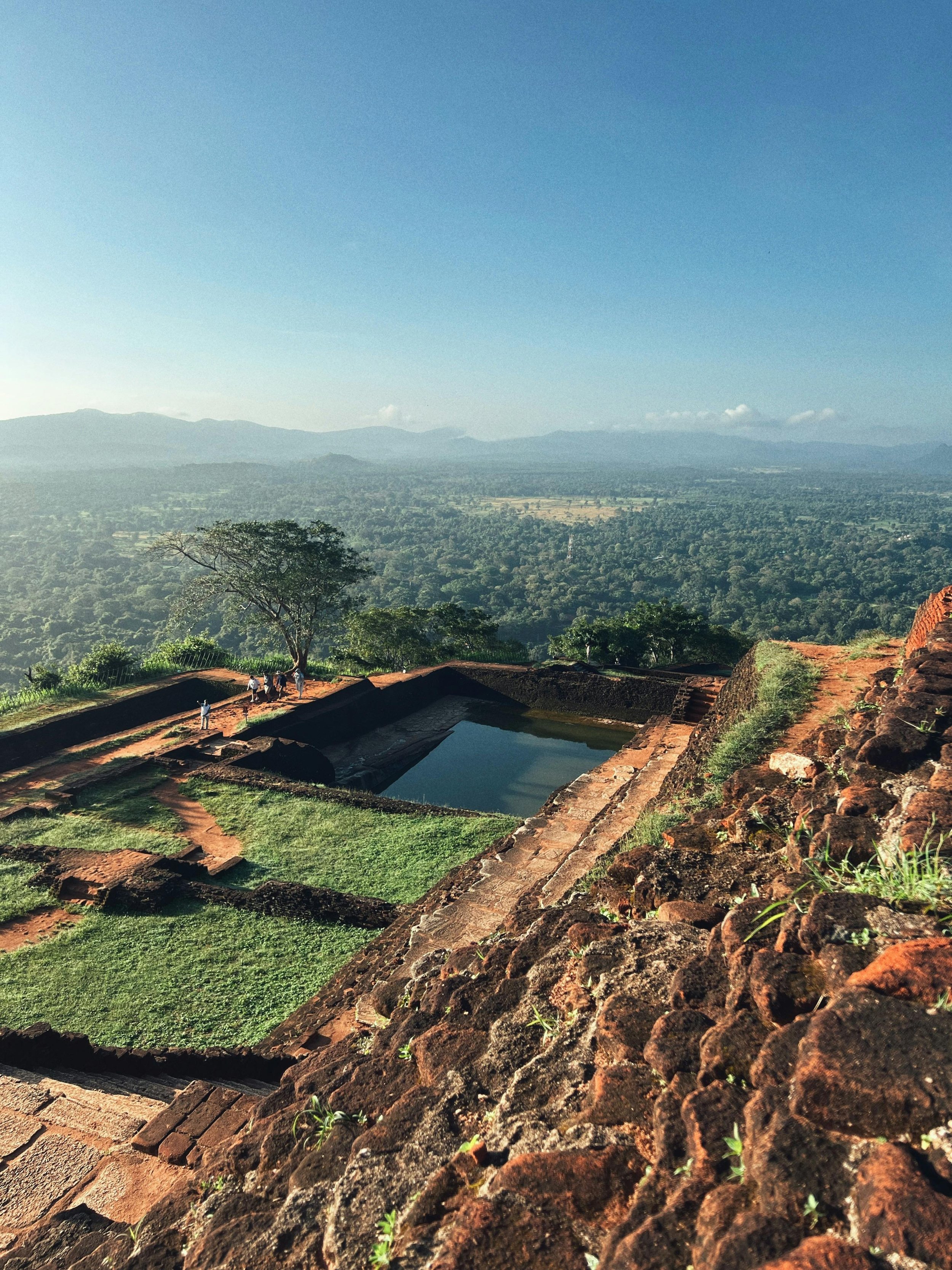 Ancient stone ruins with a pool, surrounded by greenery, overlooking a valley with trees and mountains in the distance under a clear blue sky.