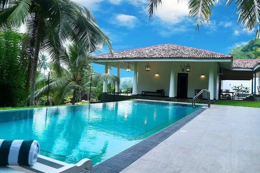 Luxury poolside scene with a modern house, lush green tropical trees, and a bright blue sky.