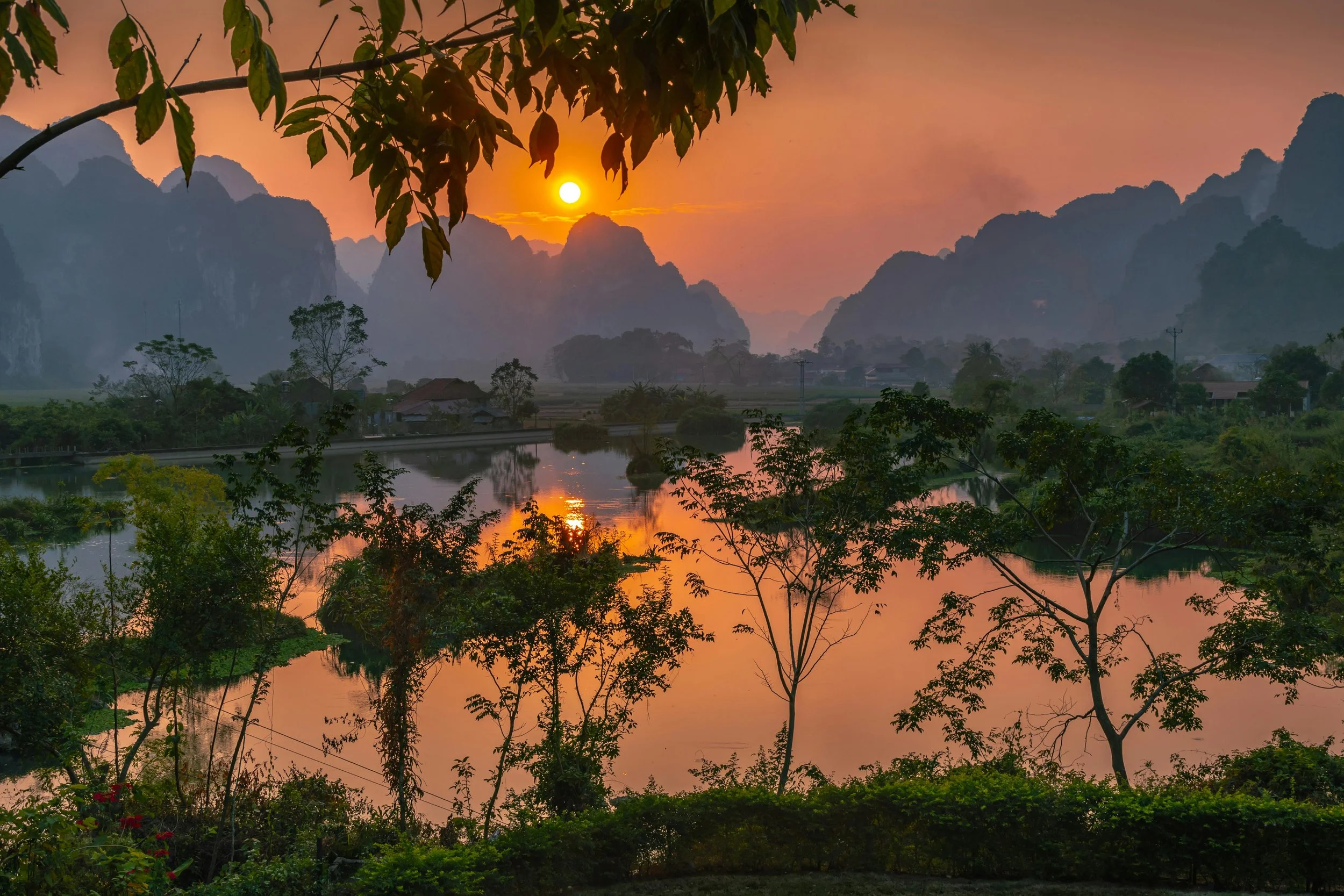 Sunset over a river with mountains in the background and trees in the foreground.