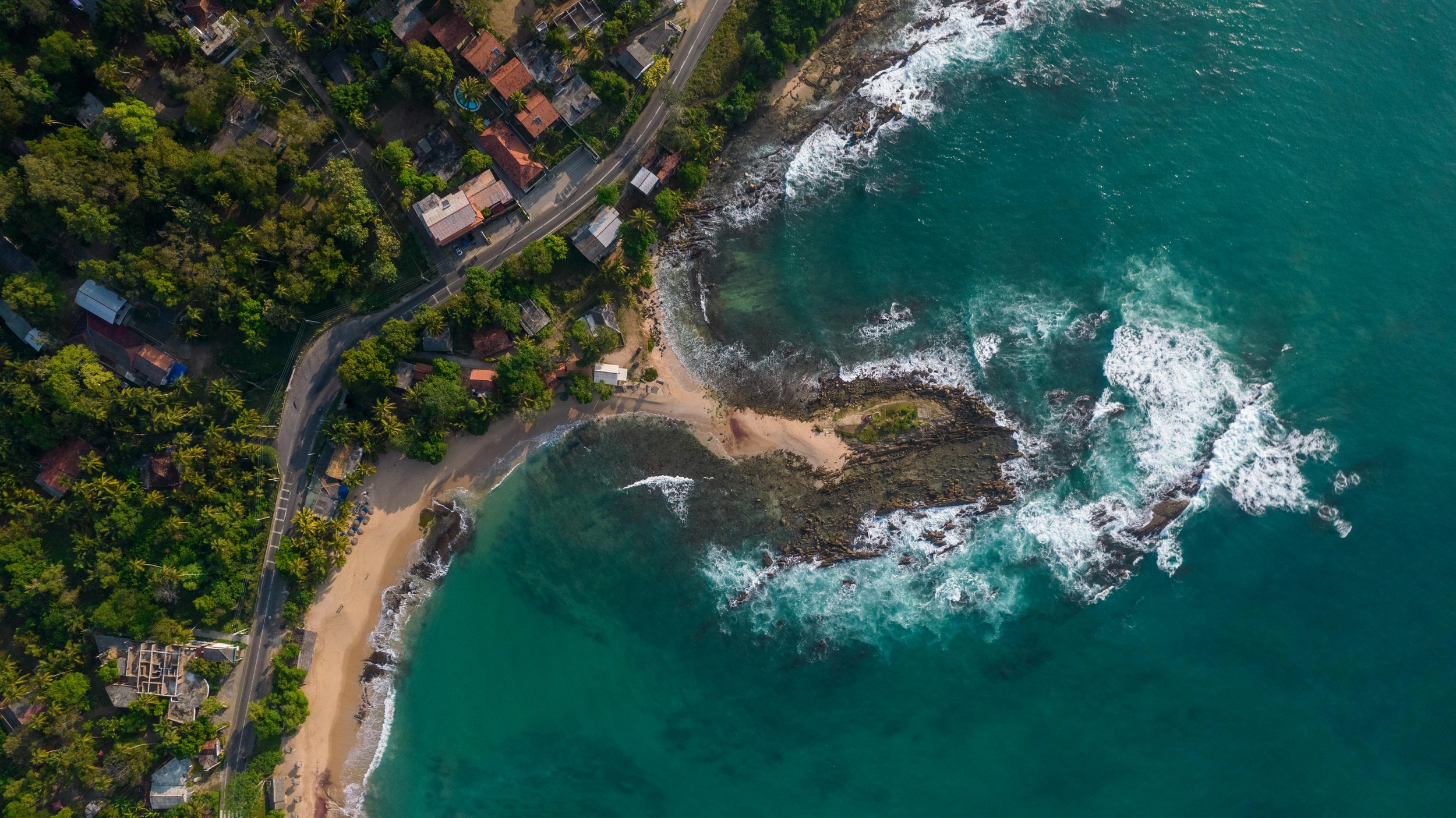 An aerial view of a coastline with turquoise water, rocky shoreline, small beach, and lush green trees. There are houses and buildings along a winding road near the forest.