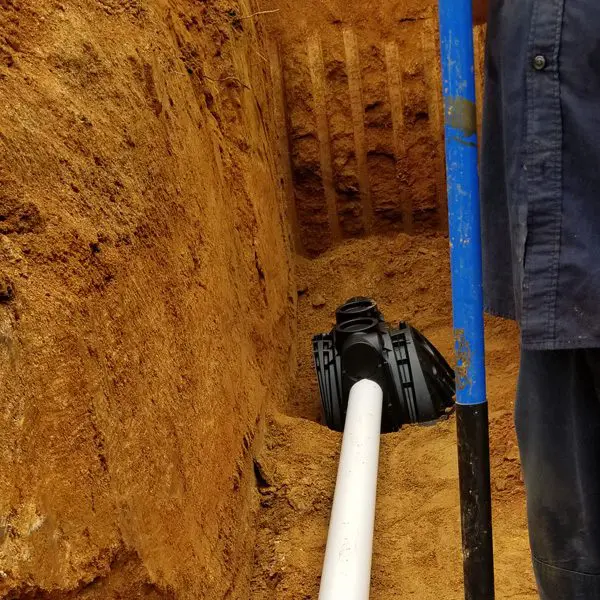 A trench with a black drainage or sewer pipe and a white PVC pipe, with a person standing nearby holding a blue-handled tool.