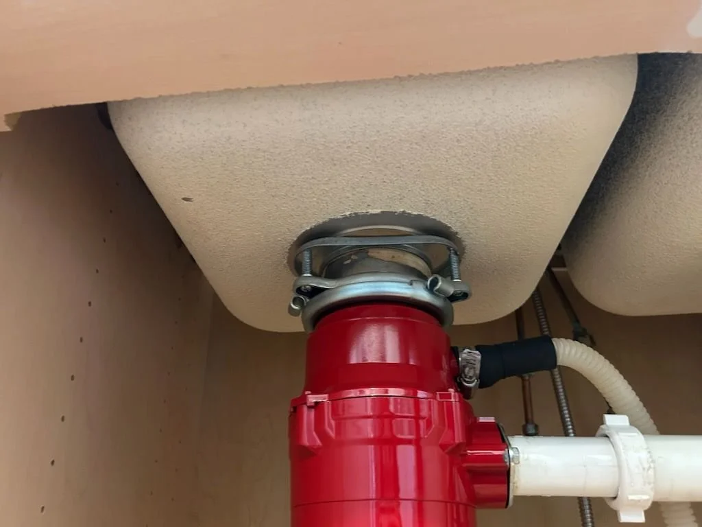 Underneath a kitchen sink showing the drain pipe connected to the sink with a rubber seal and metal clamp, along with PVC pipes and flexible hoses.
