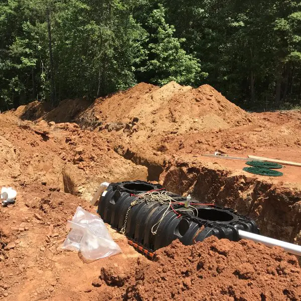 Underground septic tank installation site with black septic tanks partially buried in a dug trench, surrounded by piles of excavated red soil, with some green garden hose coiled in the background and a white PVC pipe visible in the trench.