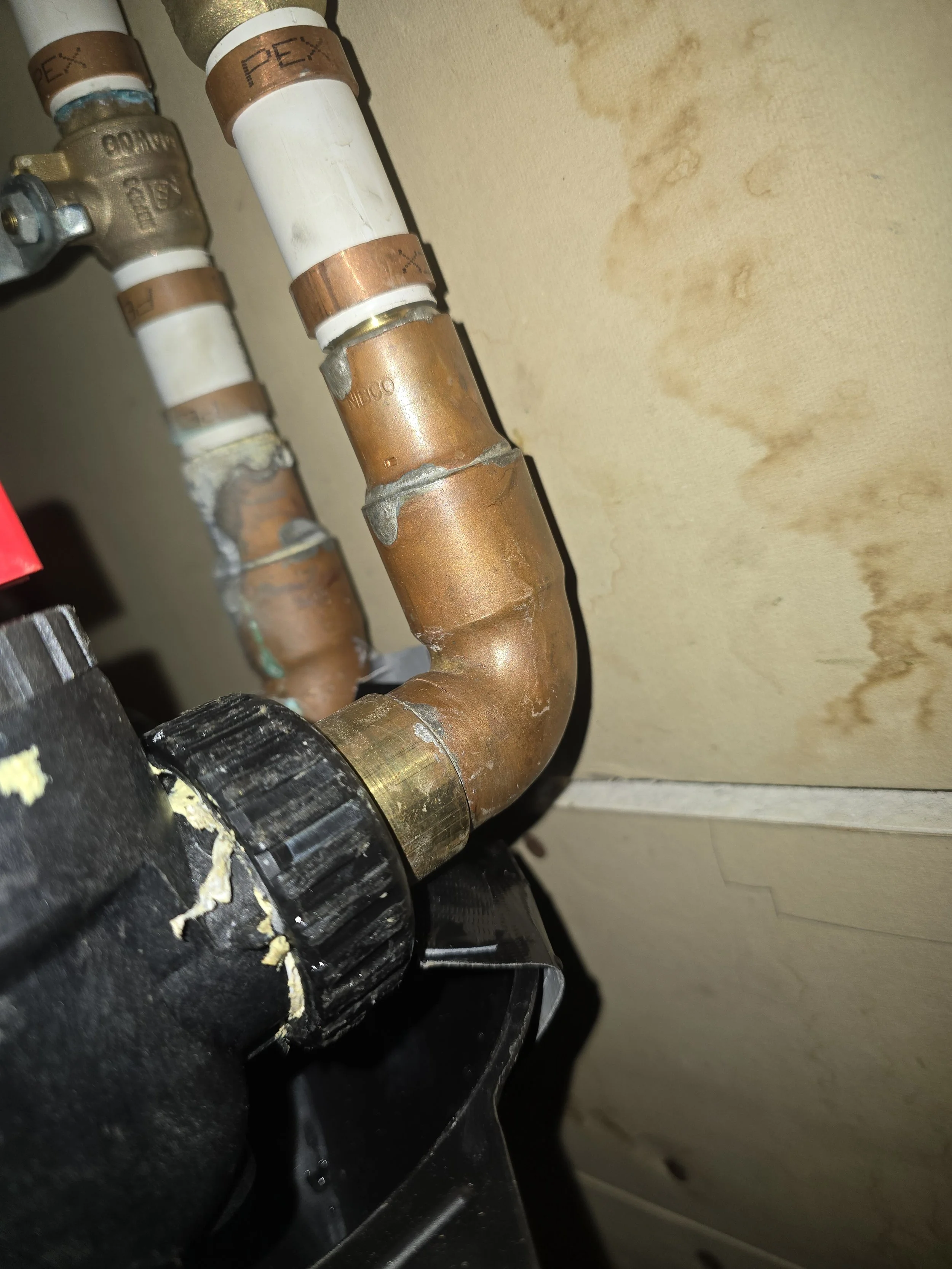 Close-up view of copper and plastic pipes with fittings, connected to a valve at the top, against a light-colored wall.