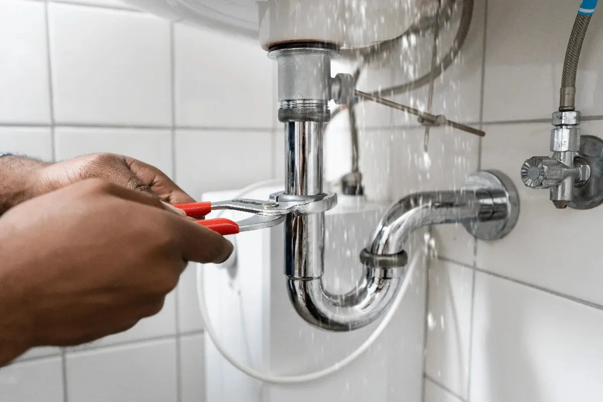 A person using pliers to tighten a pipe under a sink.