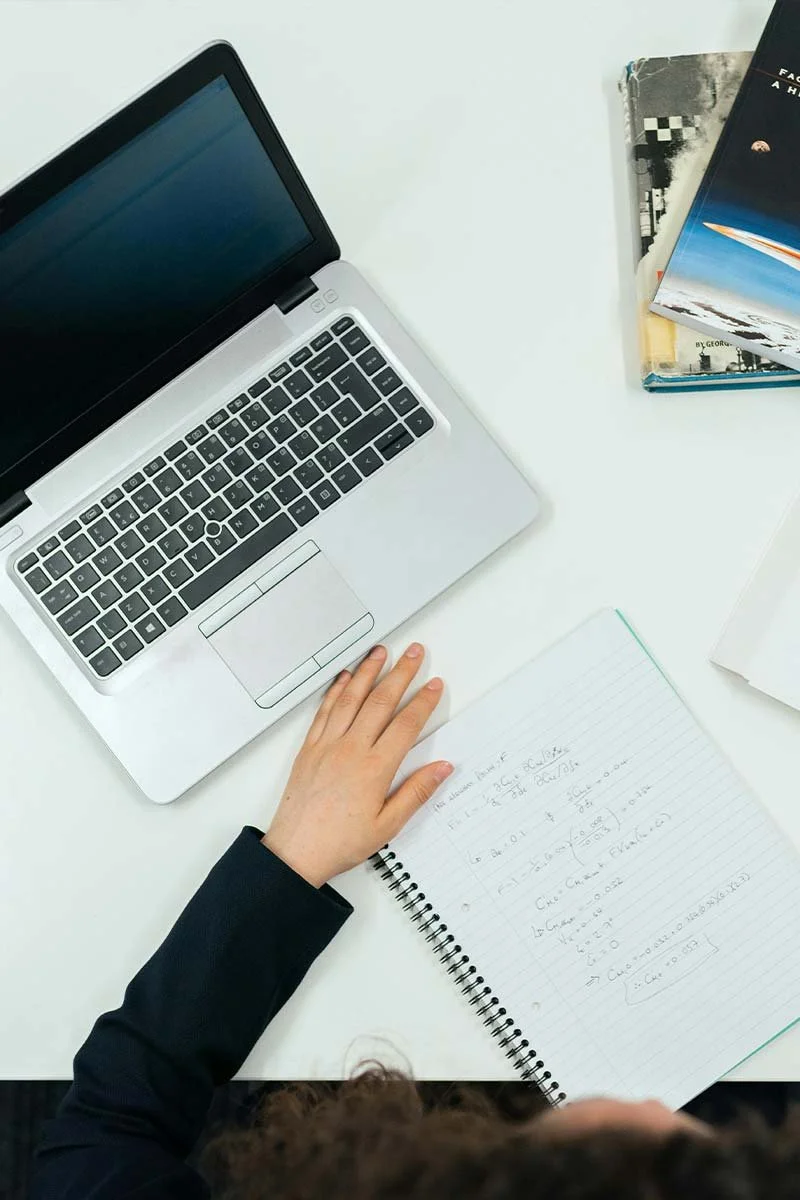 Overhead view of a student working at a white desk with a laptop, notebook, textbooks, and papers.