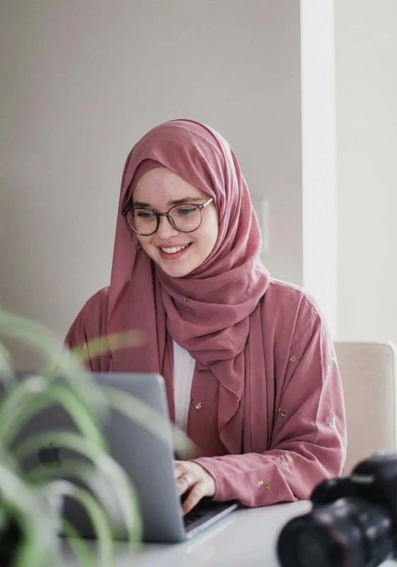 Young woman wearing a pink hijab and glasses, smiling while working on a laptop at a desk.