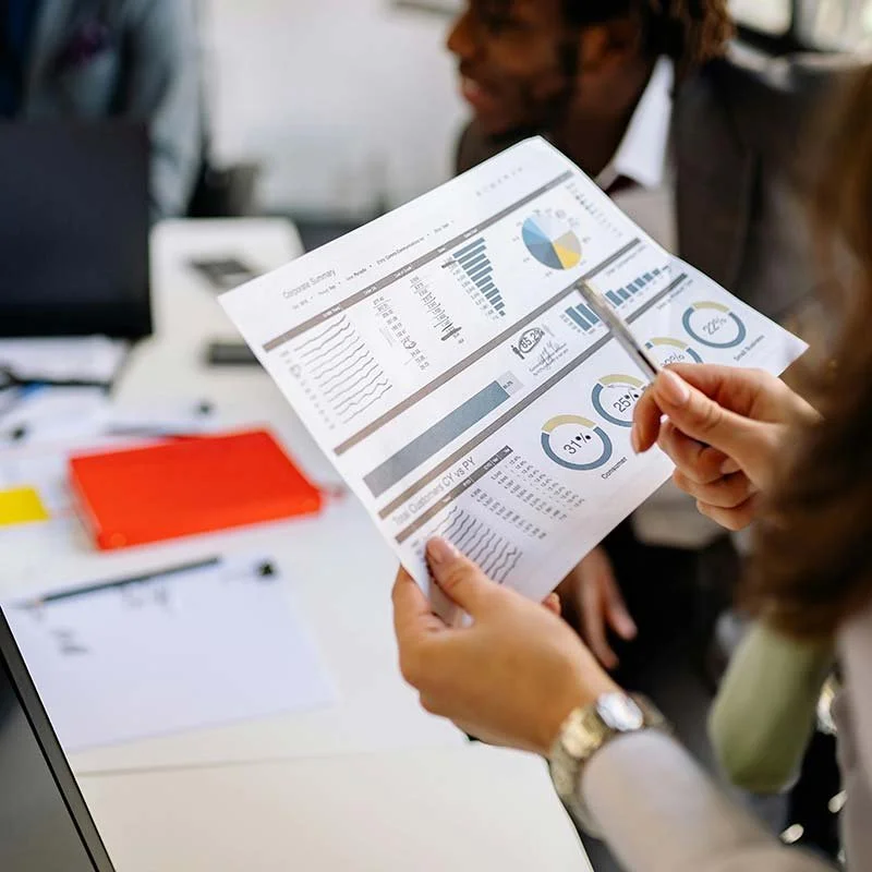 Person holding a paper with financial charts and graphs, pointing at it with a pen, in a business meeting.