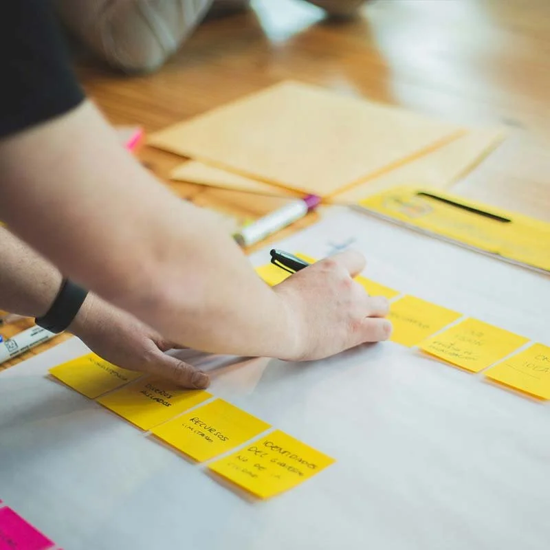 Person organizing yellow sticky notes on a table, with some orange folders and a white marker visible in the background.