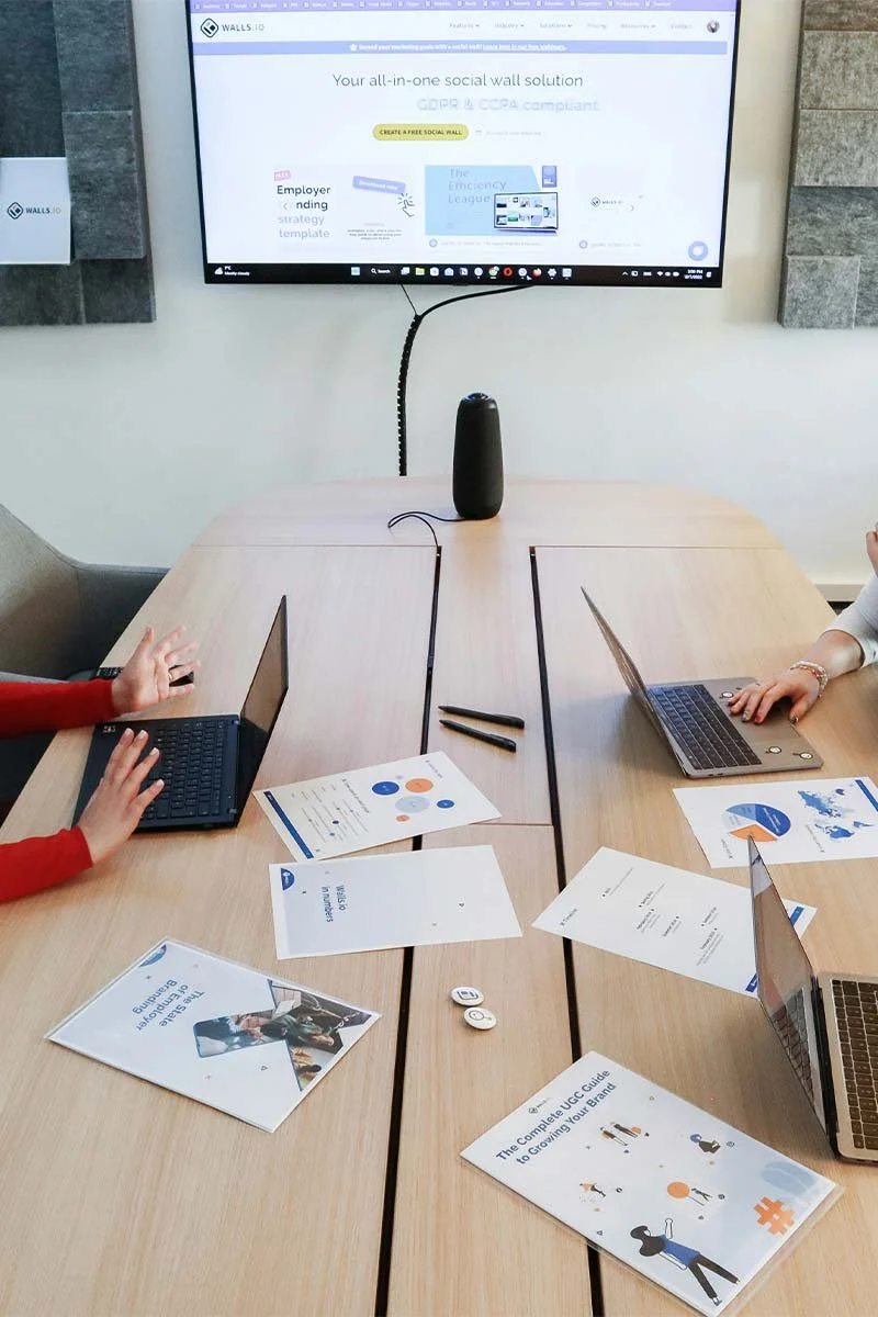 Meeting room with a wooden conference table, two laptops, project materials, and a large monitor displaying a website about social wall solutions.