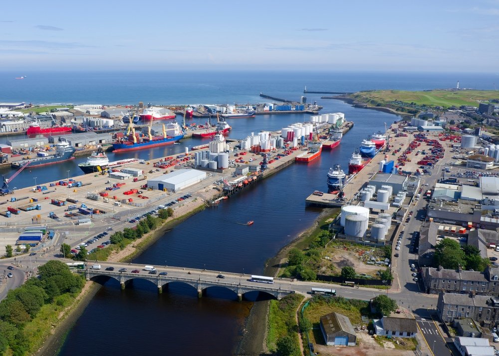 Aerial view of a busy port with ships docked, storage tanks, industrial buildings, a bridge, and the ocean in the background.