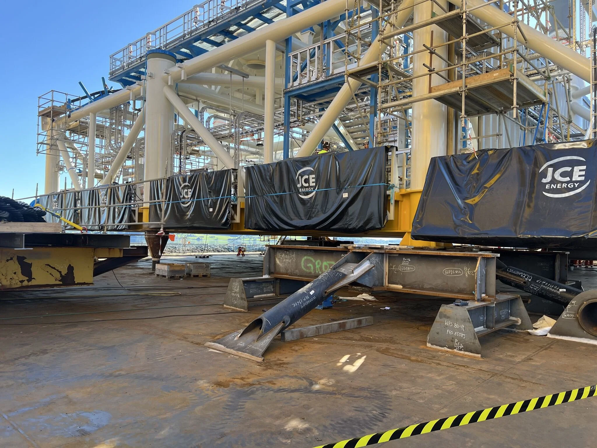Construction site with large industrial equipment, scaffolding, and metal framework, with black protective coverings labeled 'JCE Energy' in the foreground, and a clear blue sky in the background.