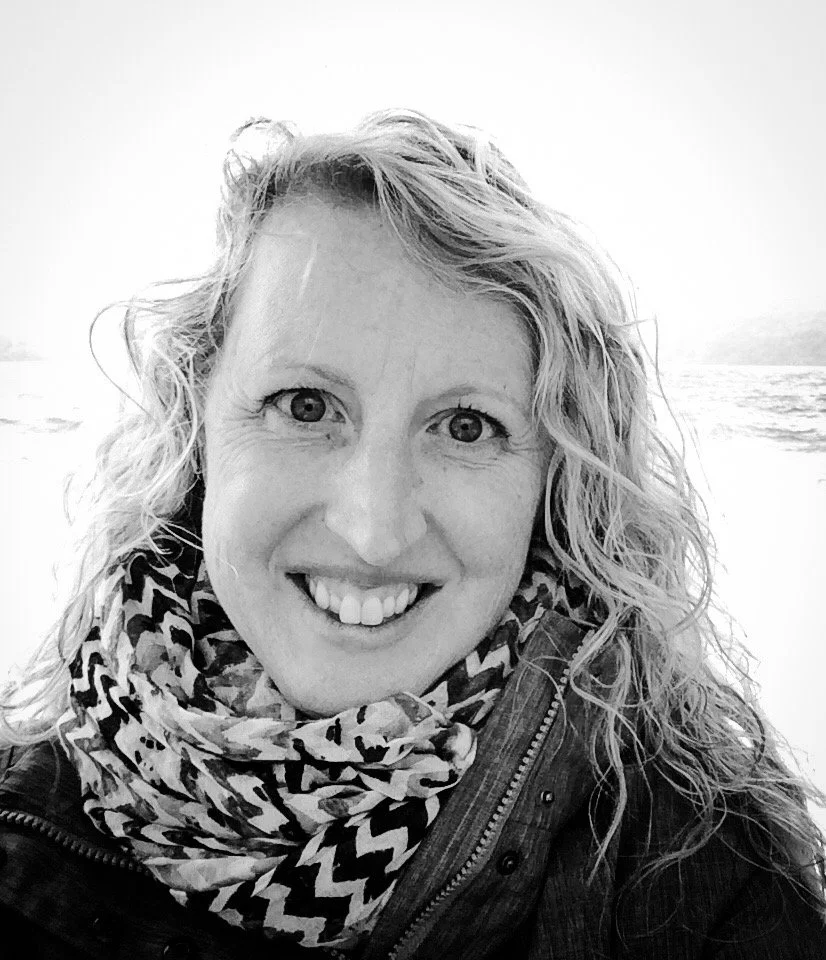 A smiling woman with curly hair wearing a patterned scarf and jacket, outdoors near water.