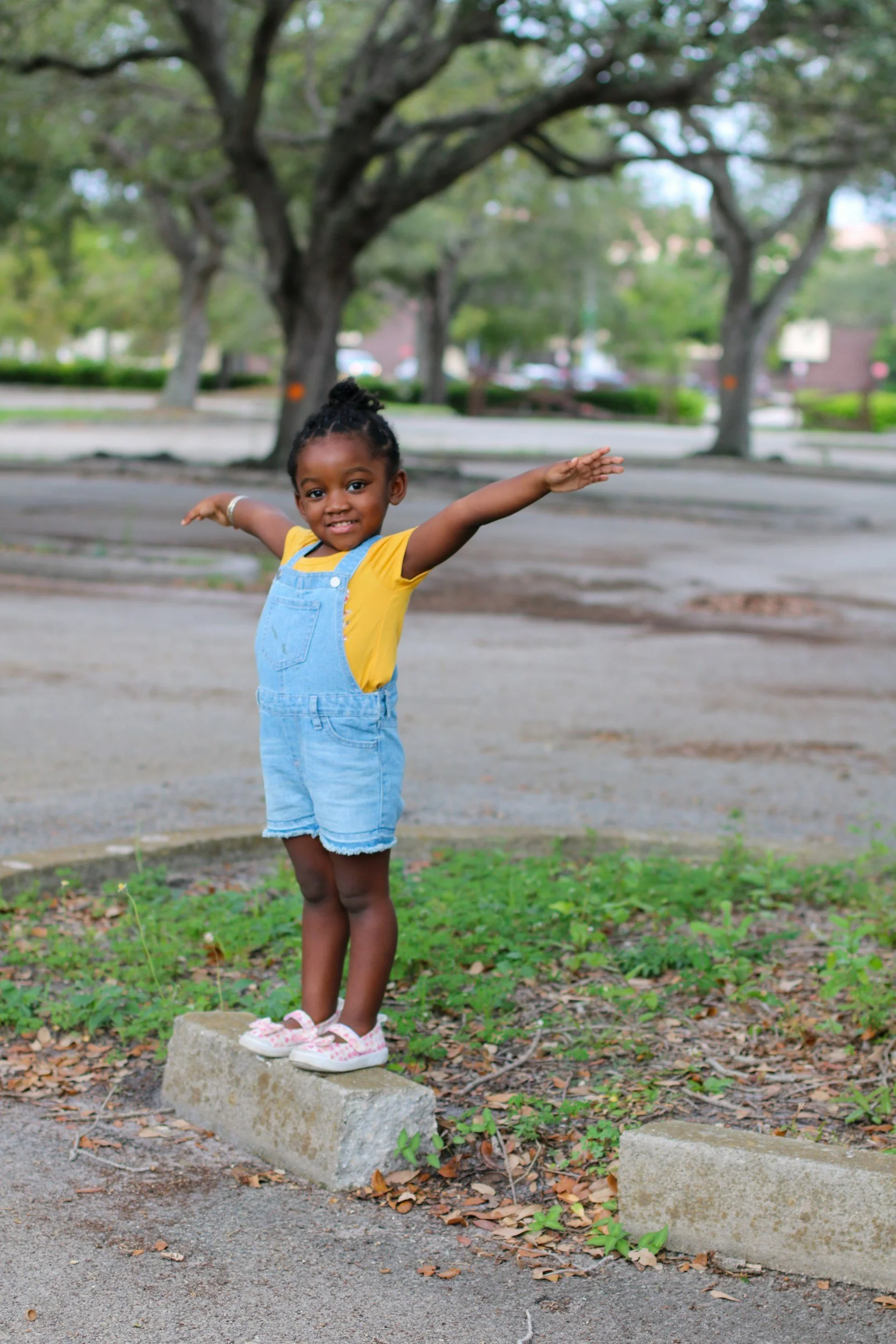 A young girl standing on a curb with arms outstretched in a park, smiling, with trees and a parking lot in the background.
