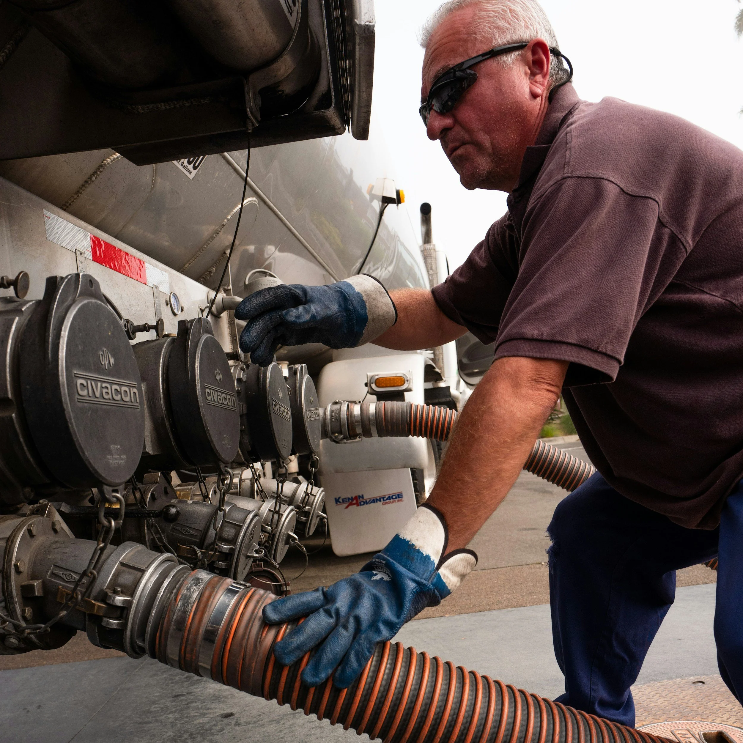 A man wearing sunglasses, gloves, and a brown shirt is connecting a flexible hose to a large industrial machine or fuel tank.