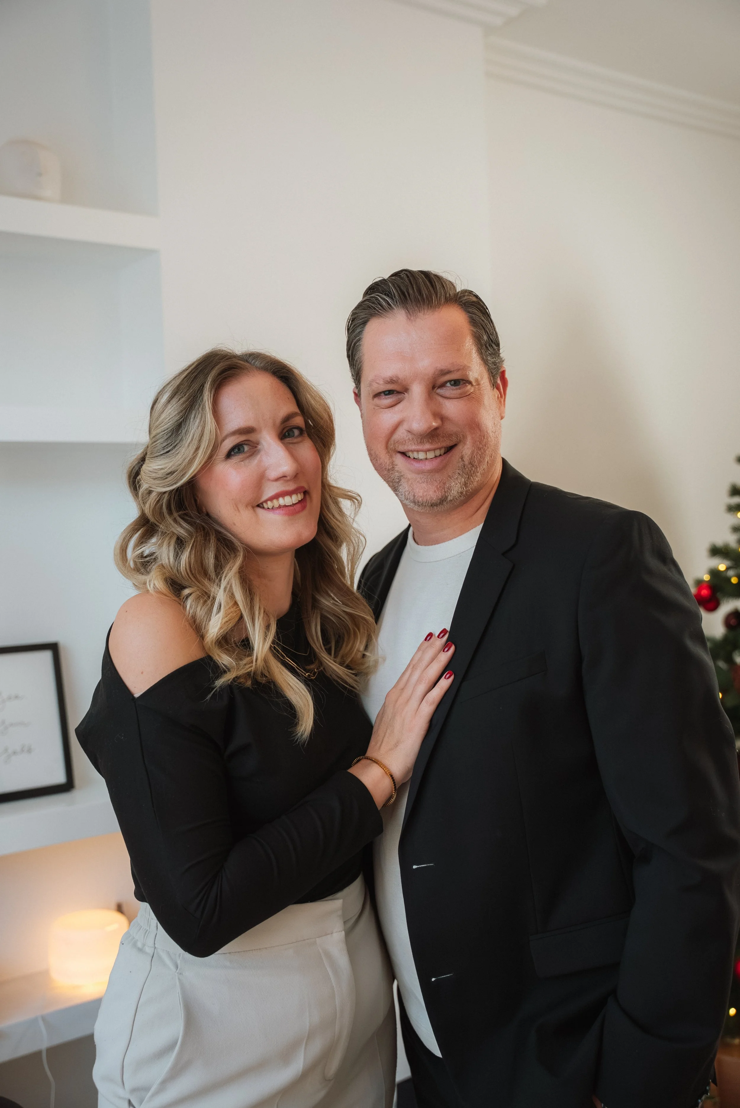 Een gelukkig stel poseert dicht bij elkaar in een huis, met een achtergrond van een kerstboom met rood en gouden ornamenten.