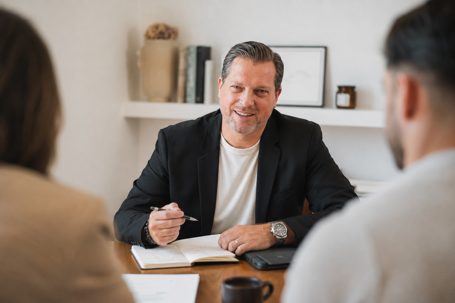 Man in zakelijk jack en wit shirt die glimlacht tijdens een gesprek aan tafel met twee andere mensen.