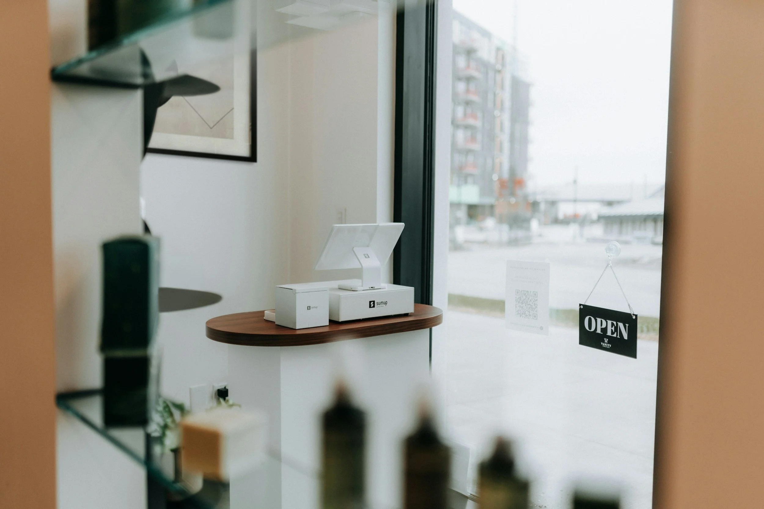 Binnenkant van een winkel met een tafel waarop een apparaat staat en een 'Open' bordje aan de deur met ramen in de achtergrond.