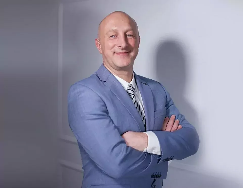 Man in een blauwe pak met een wit overhemd en gestreepte stropdas, hij staat met gekruiste armen en lacht.