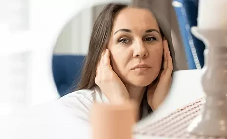 Vrouw die in de spiegel kijkt met een hand op haar gezicht, mogelijk in een medische of klinische omgeving.