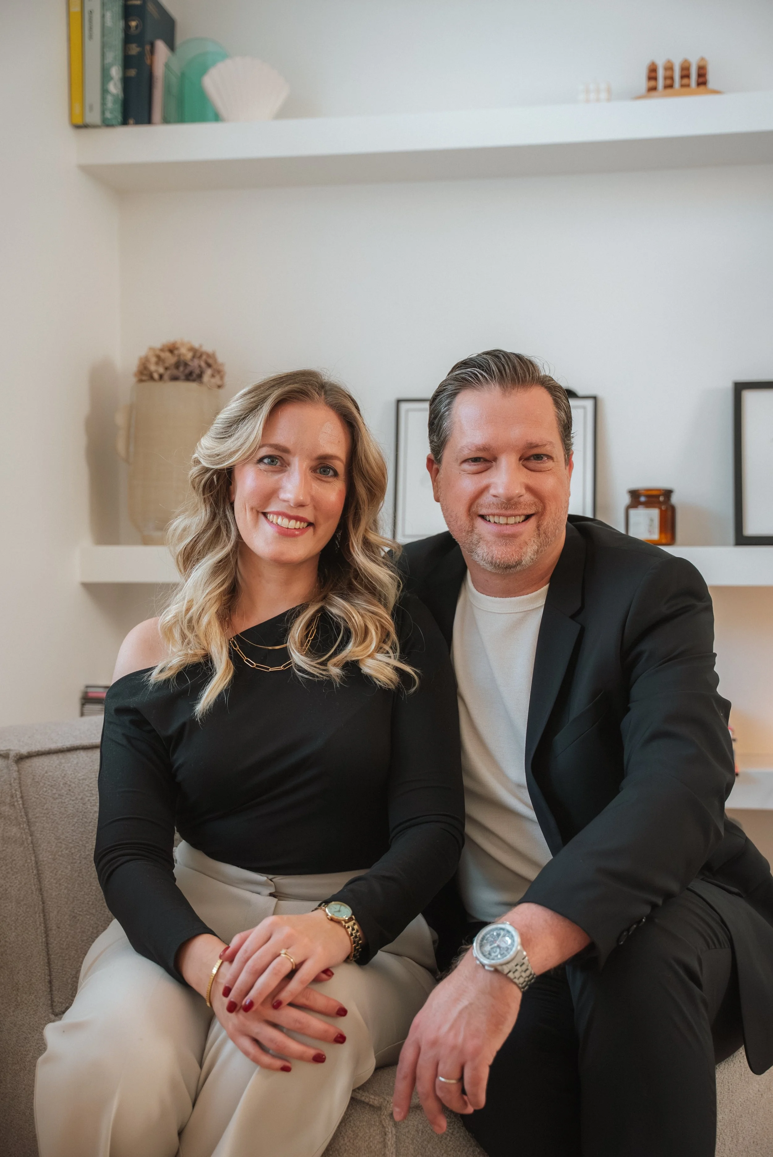 Een man en een vrouw zitten dicht naast elkaar op een bank, glimlachen naar de camera, in een huiselijke omgeving met planken en decoraties op de achtergrond.